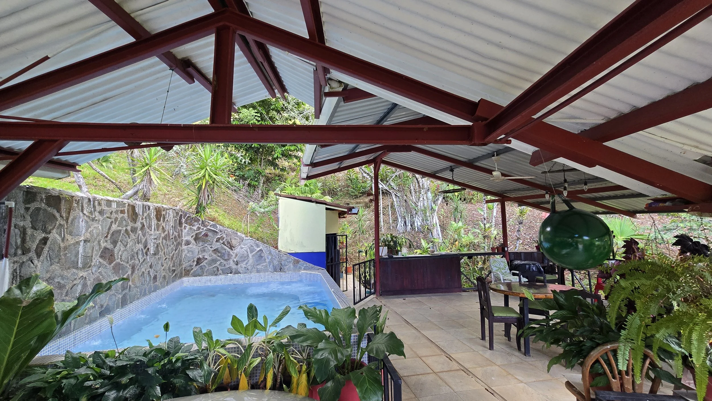 Outdoor covered patio with hot tub, plants, tables, chairs, and a green hanging ball, surrounded by a hillside with trees and greenery.
