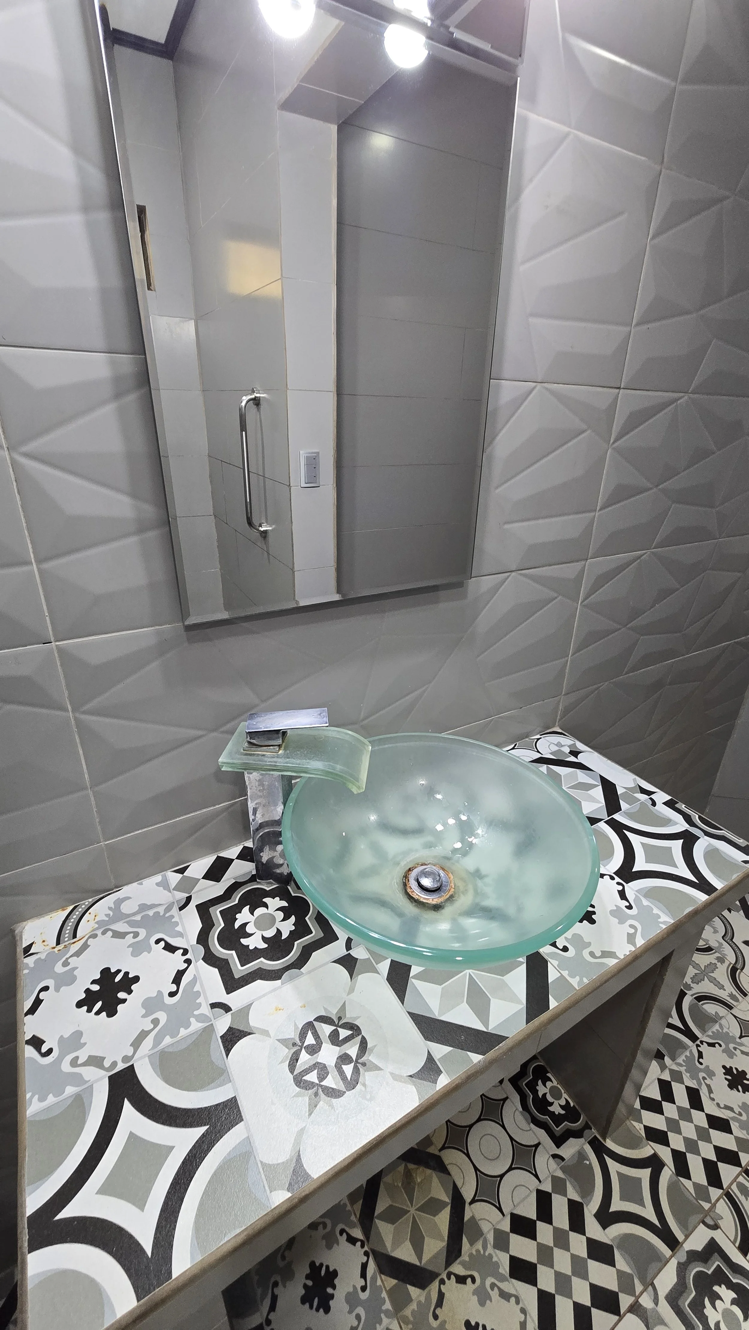 A bathroom vanity with a decorative black and white patterned tile countertop, a round glass vessel sink, and a modern faucet; a mirror behind the sink reflects a door, handle, and light fixtures.