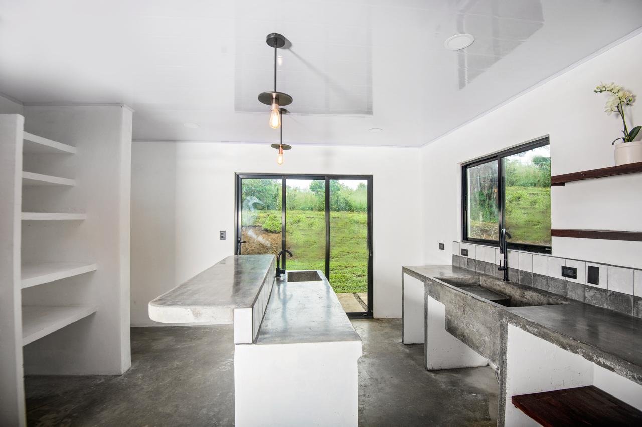 Modern kitchen with white walls, concrete countertops, black sinks, and open shelving, overlooking a grassy outdoor area through sliding glass doors and a window.