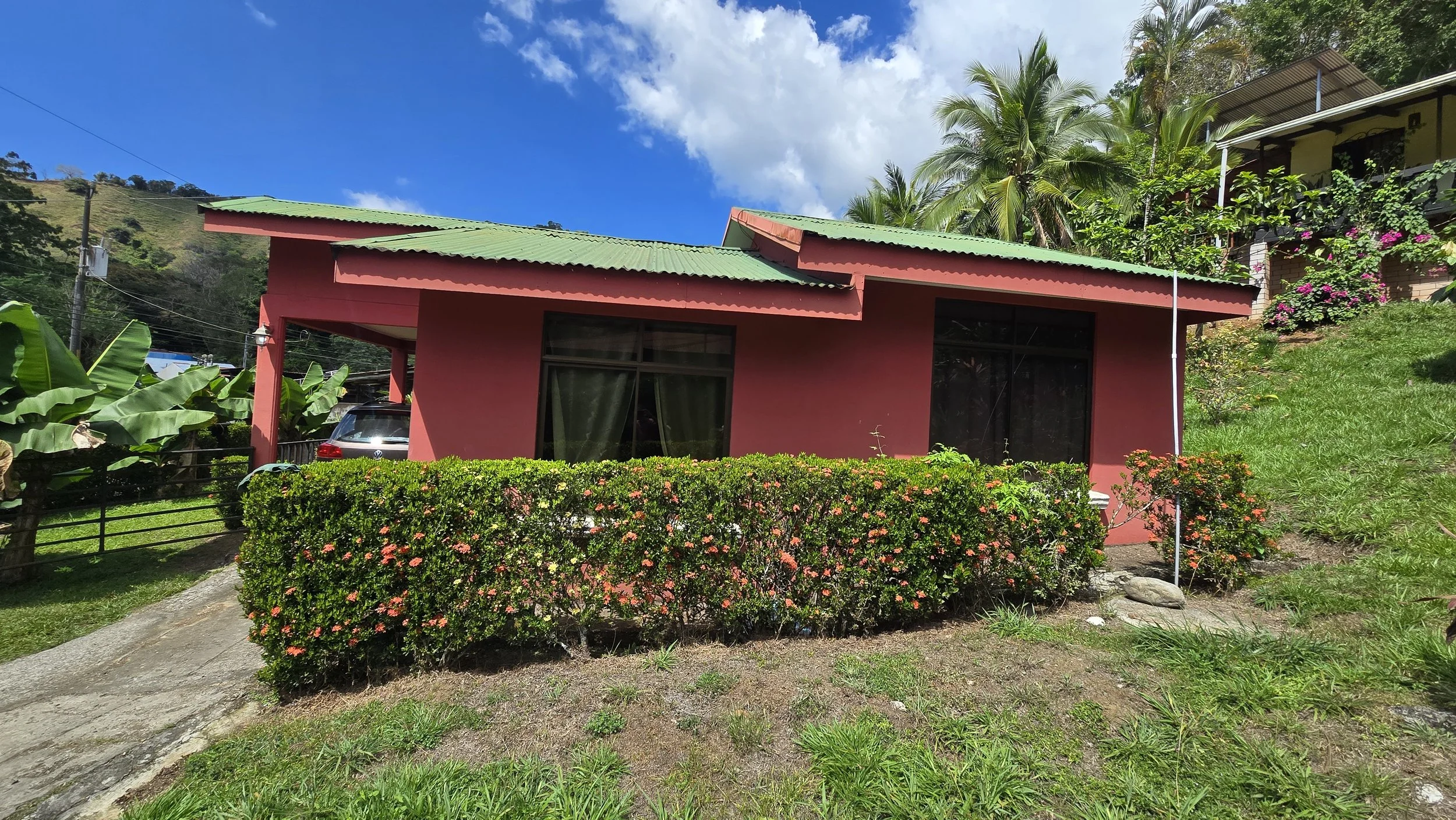 A pink house with a green roof, surrounded by lush greenery and tropical plants, including banana trees and palm trees, under a partly cloudy blue sky.
