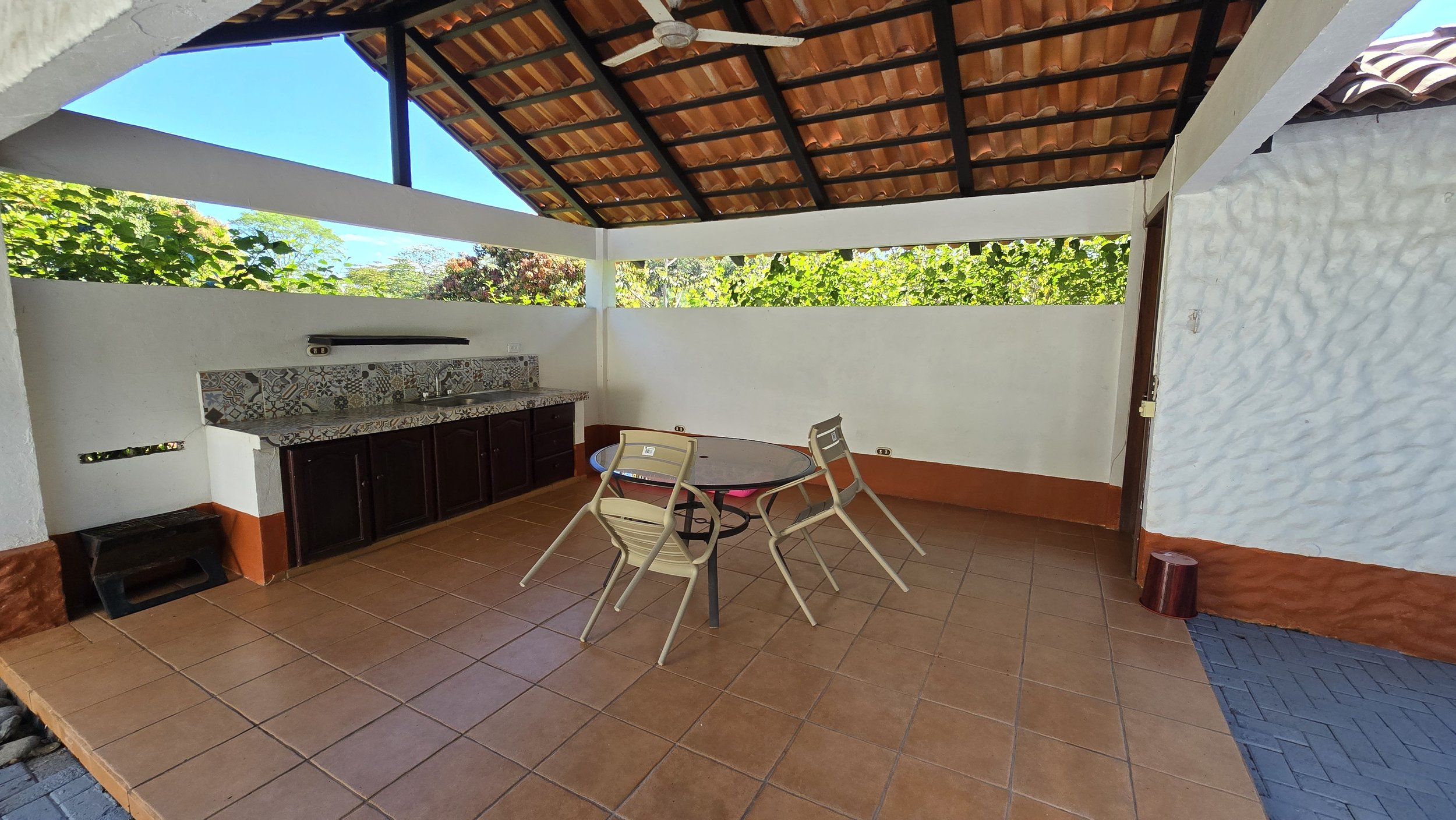 Outdoor covered patio area with ceramic tile flooring, a round glass-top table surrounded by four plastic chairs, a small dark stool, a built-in sink area with patterned tiles, and a white wall with greenery visible through an open section above.