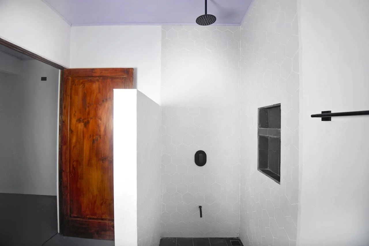 Empty bathroom with white hexagonal tile walls, black drain and niche, black showerhead, wooden door, and black towel bar.