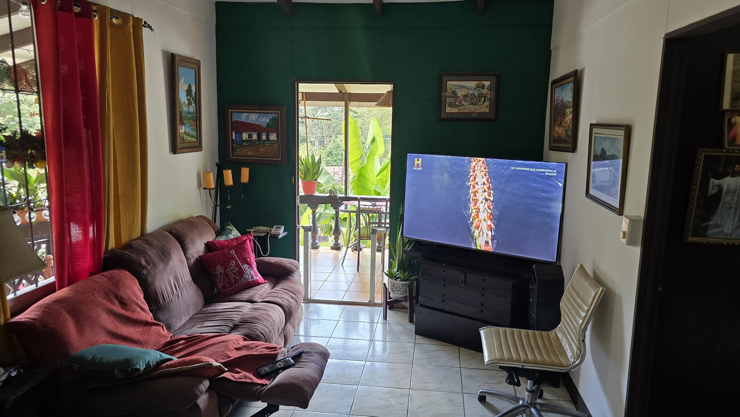 Living room with brown couch, red and green pillows, a TV on a black stand, and a small balcony with plants outside. There are paintings on the walls and colorful curtains, with a sliding glass door leading to the balcony.