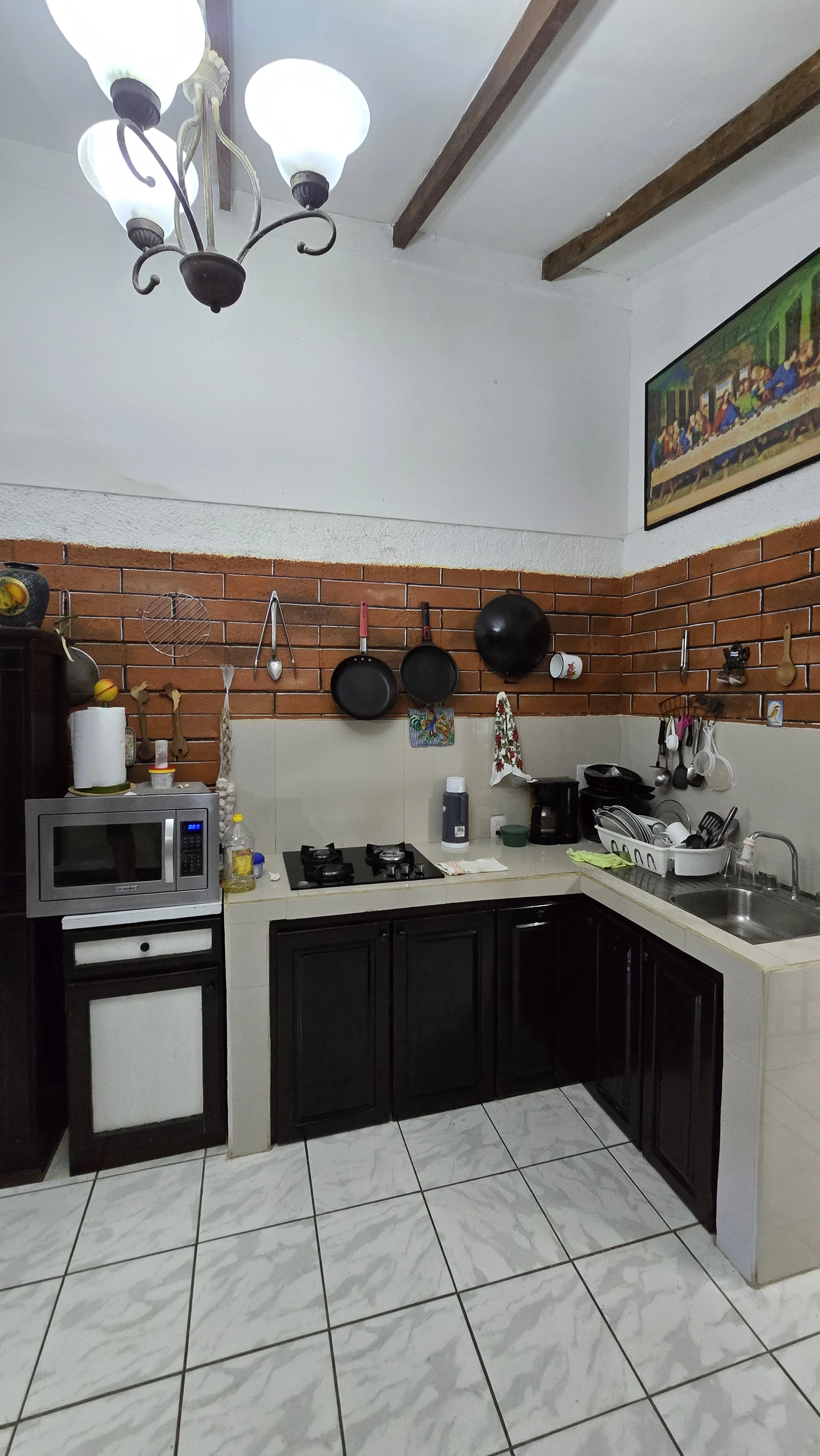 Kitchen with black cabinets, a microwave, stove, hanging pots and utensils, a sink, and a chandelier on the ceiling.