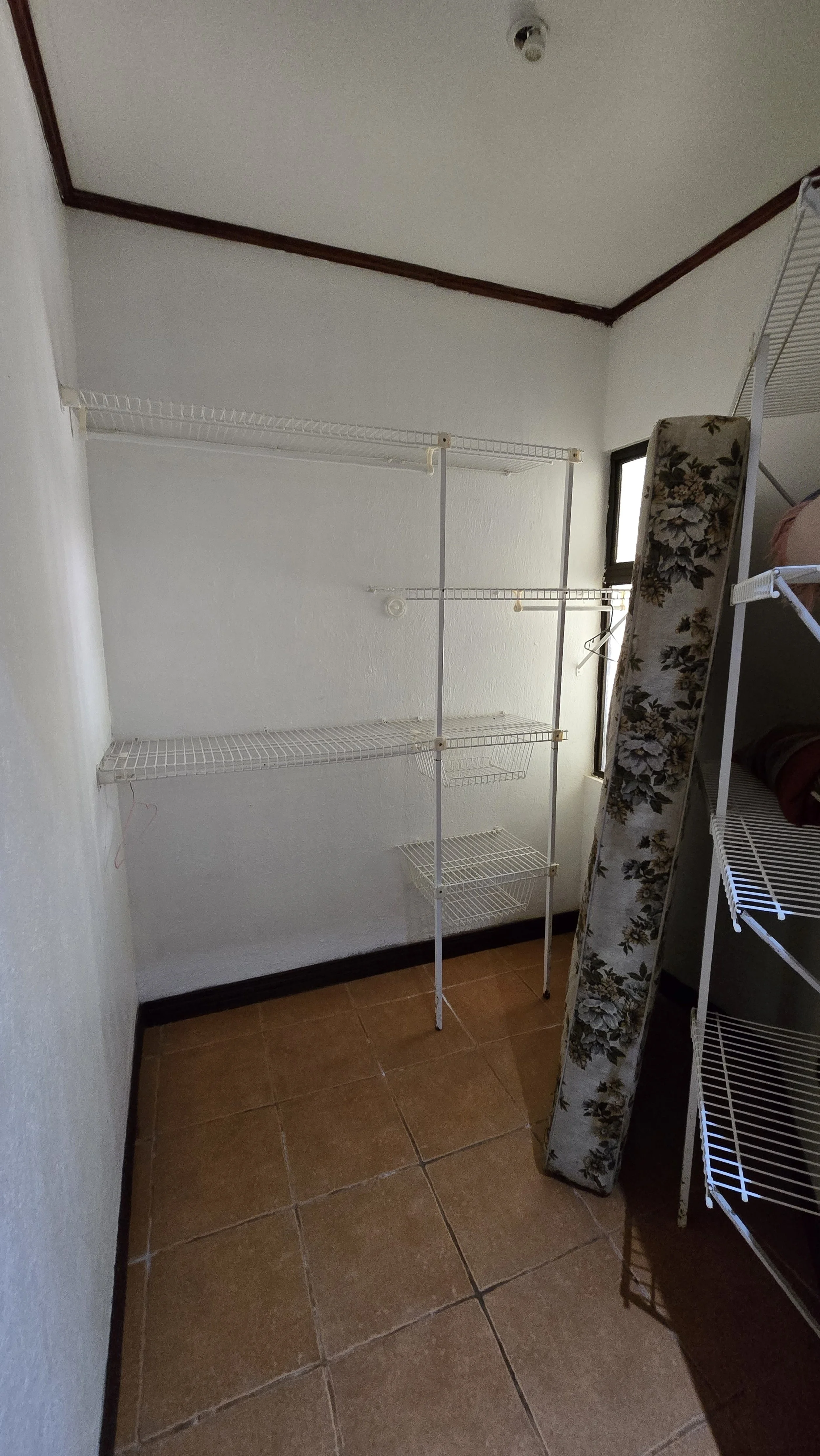 An empty wire shelving unit with four shelves is standing in a room with brown tile flooring and white walls. A window is partially visible to the right of the shelving unit, and a floral armrest is also visible to the right.