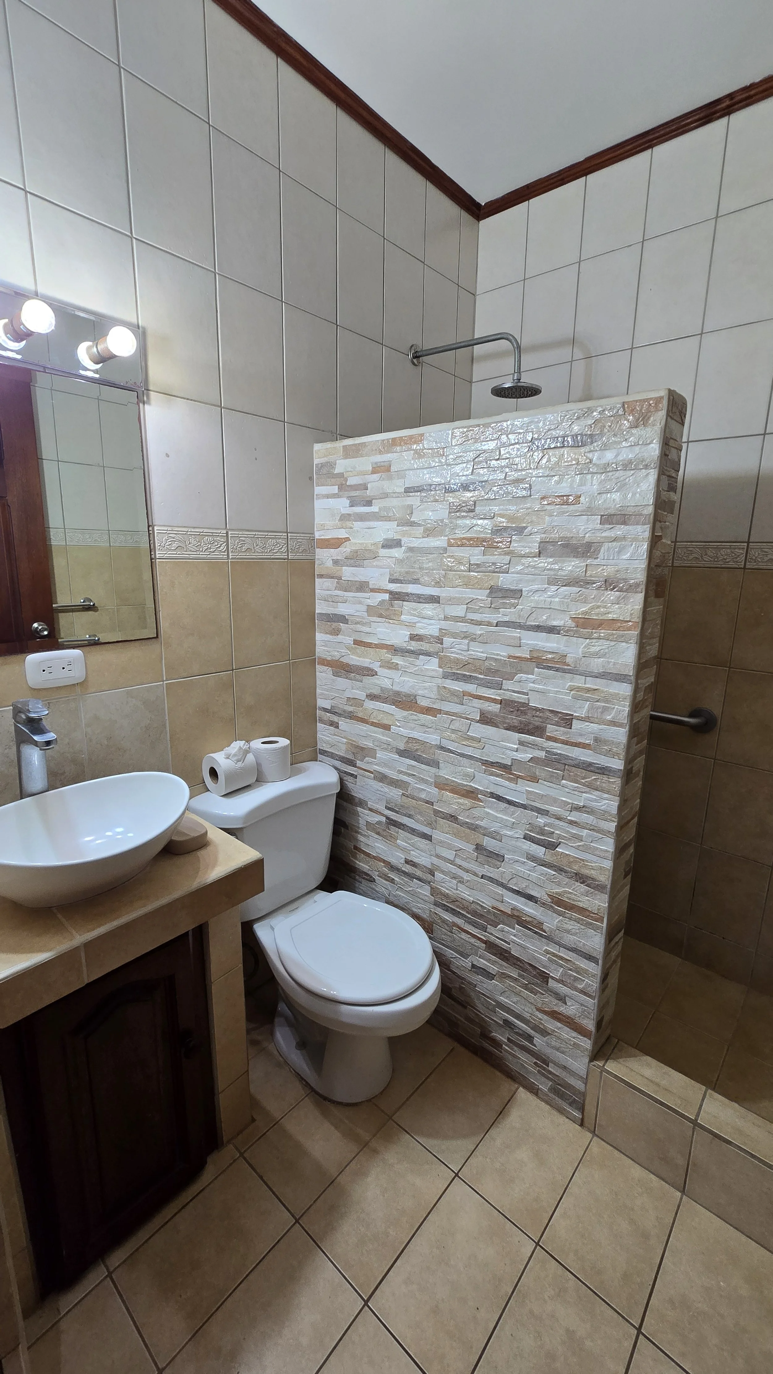 Small bathroom with beige and brown tile floors, a white toilet, a small sink with a beige countertop, a cute soap dish, a wall-mounted mirror with lights, and a shower area separated by a stone-patterned half wall.