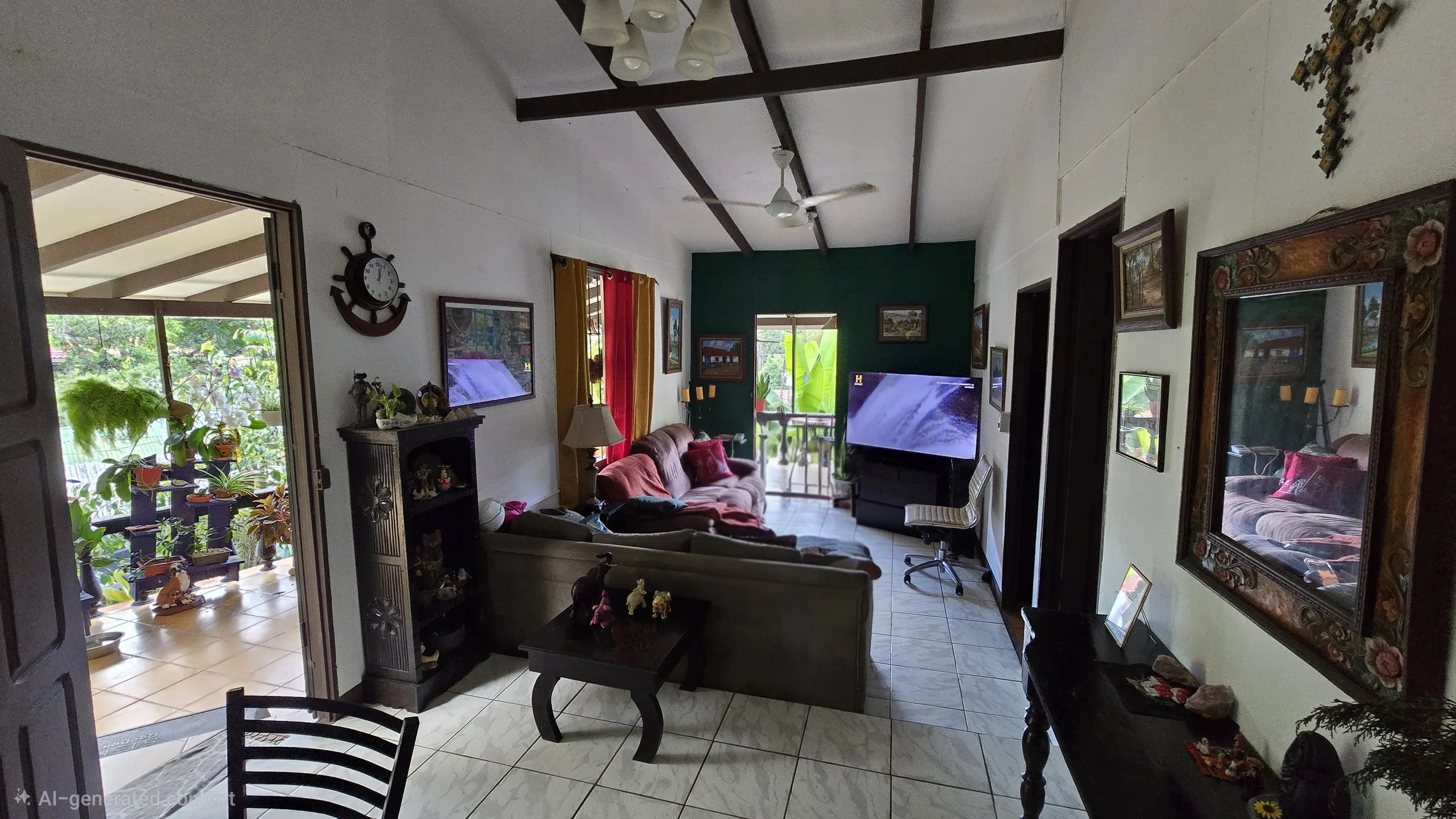 Living room with tiled floor, green accent wall, sofas, wall art, a large flat screen TV, and a balcony with tropical plants visible through the doors.