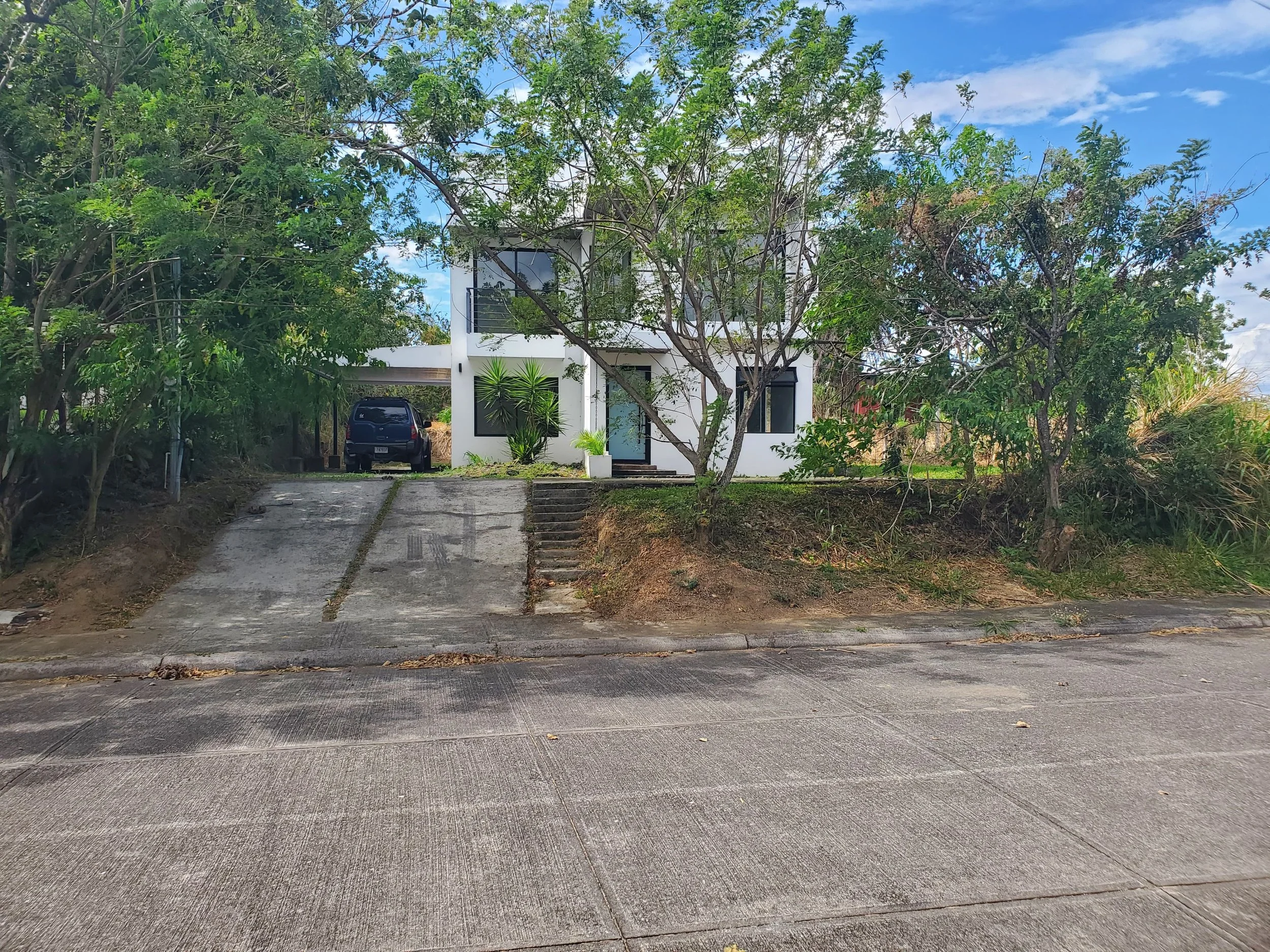 Modern white two-story house with a driveway, surrounded by trees and plants, under a partly cloudy sky.
