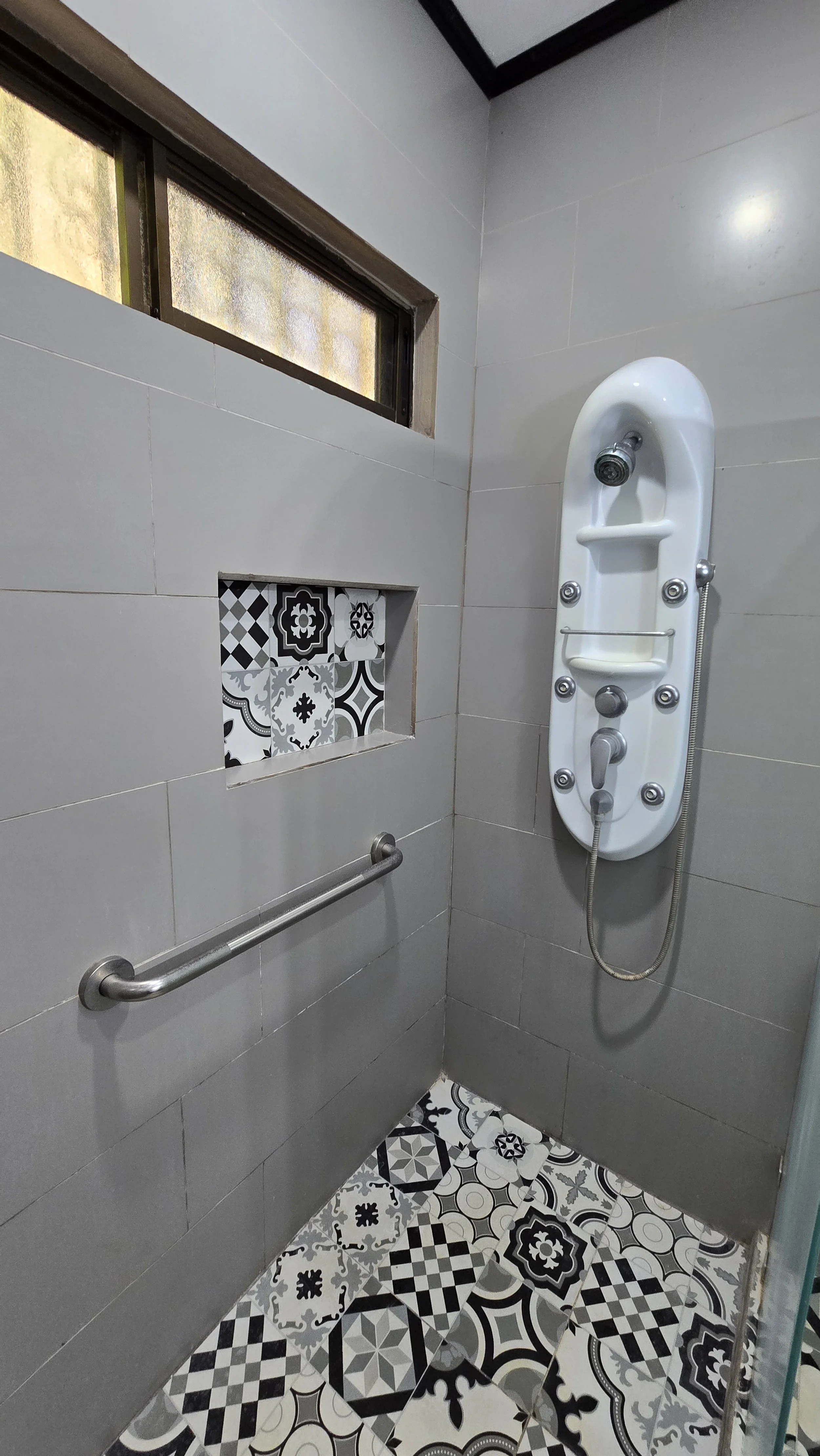 Shower stall with gray tiled walls, patterned black and white floor tiles, a horizontal grab bar, and a white shower panel with multiple jets, a handheld showerhead, and a small shelf. There is a small window at the top of the wall with frosted glass