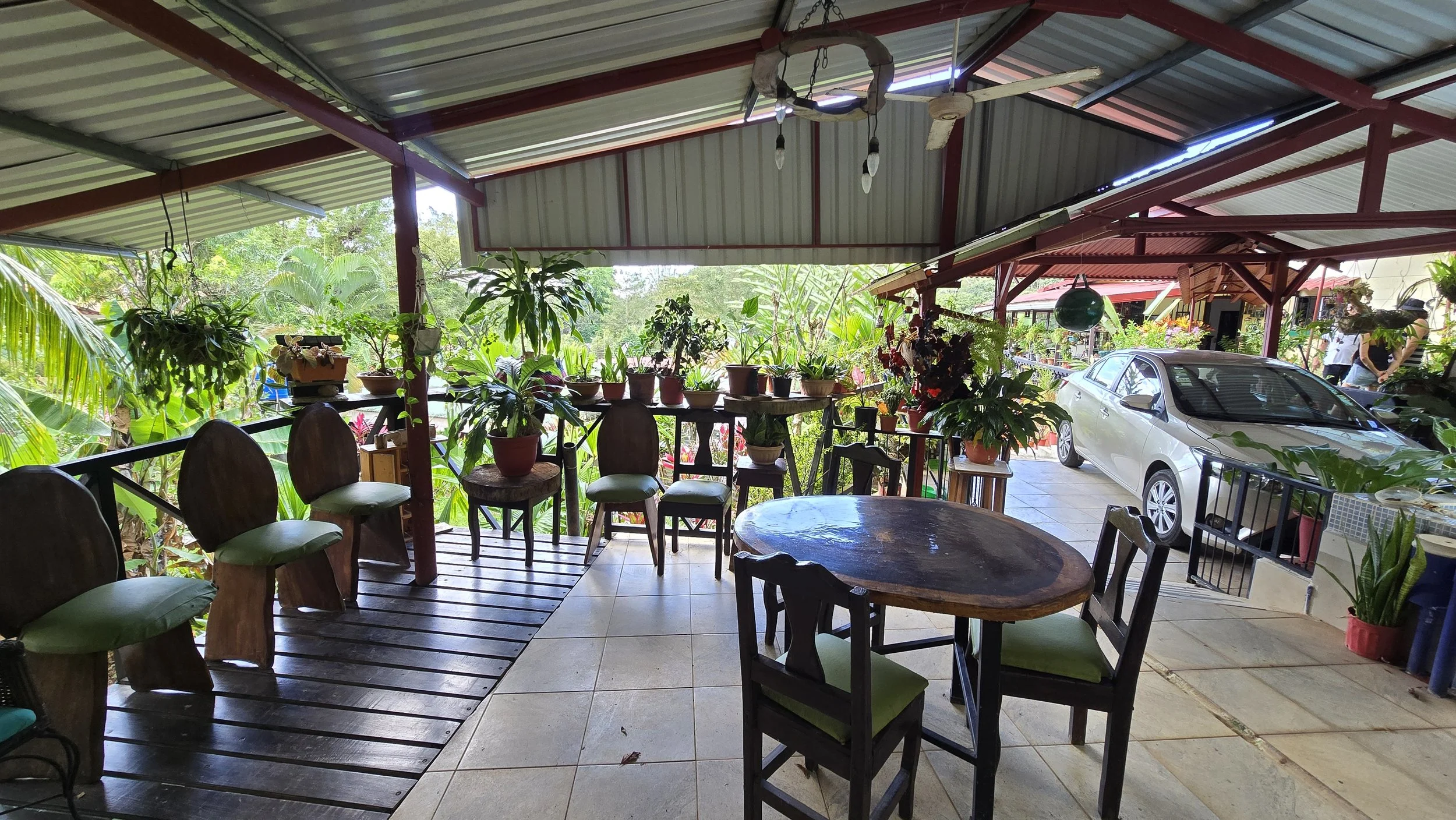Covered outdoor patio with wooden floor and tables, surrounded by potted plants and greenery, with a white car parked nearby.