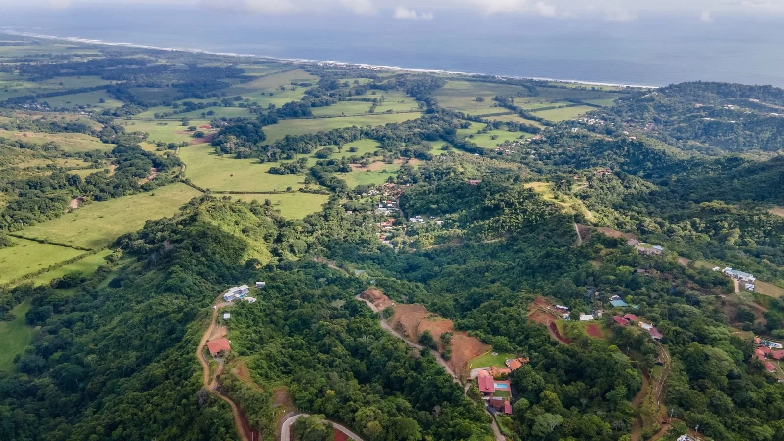 Aerial view of lush green hills, residential houses, farmland, and a coastline in the distance with the ocean meeting the sky.
