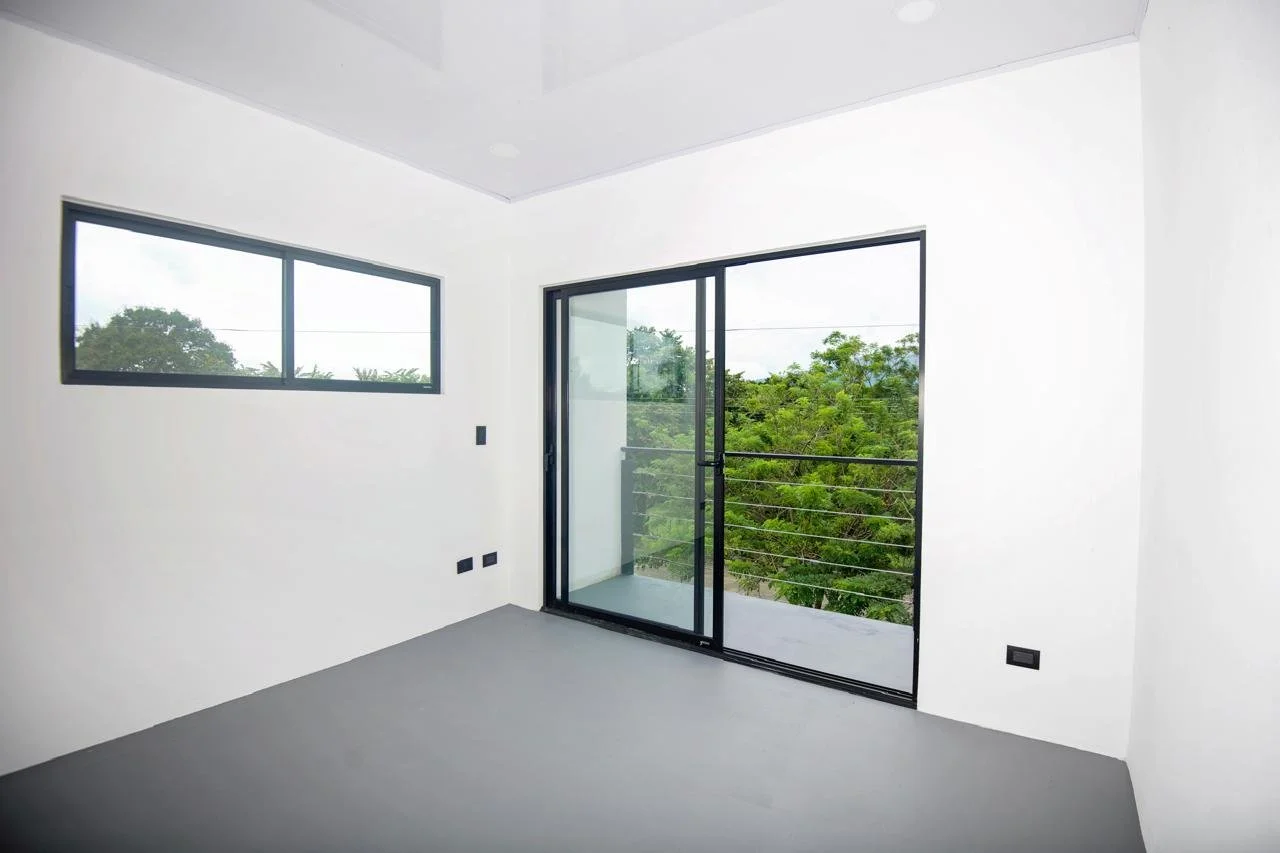 Empty white room with gray flooring, black-framed sliding glass door leading to a balcony, and black electrical outlets on the wall.