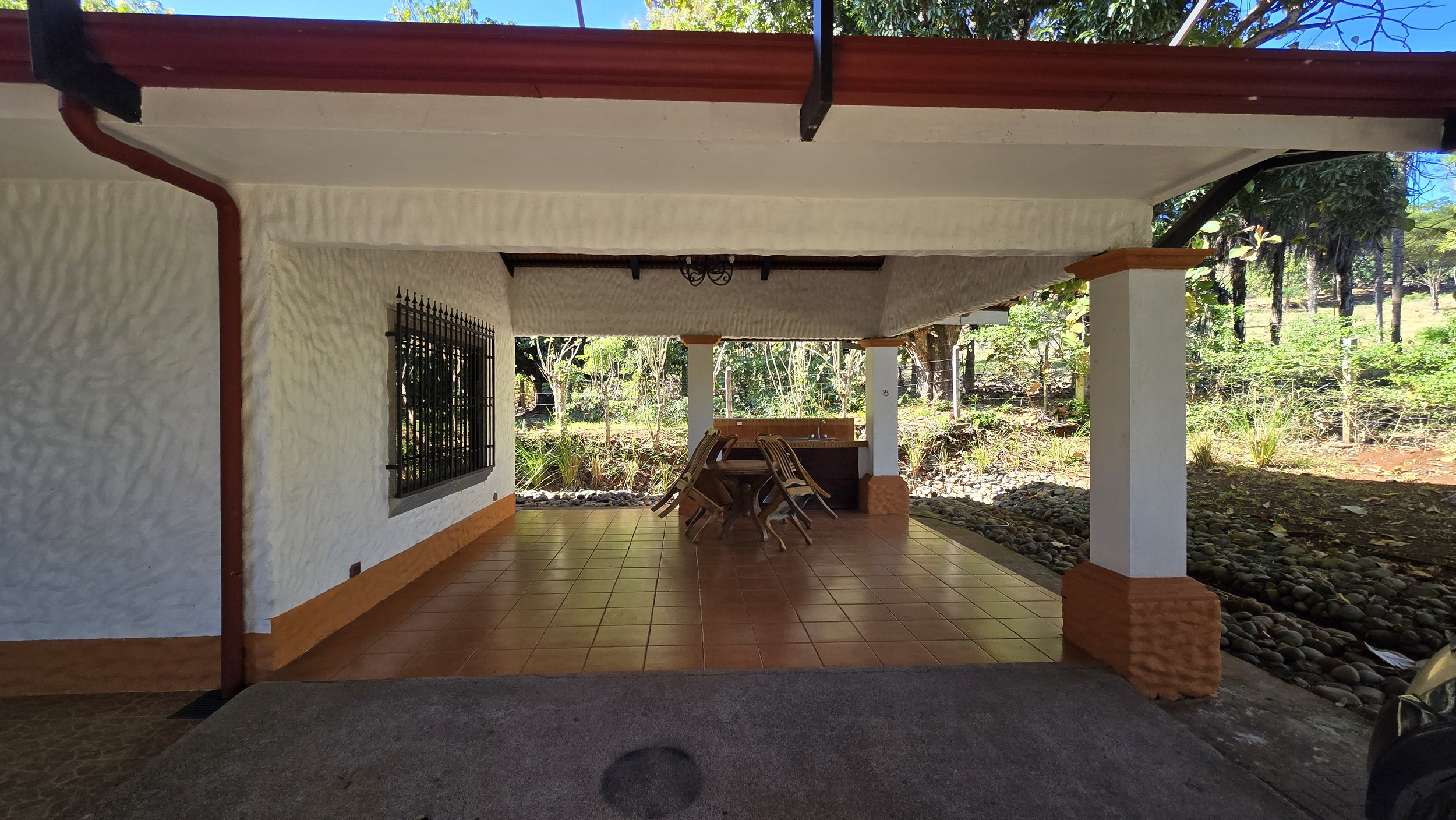 Covered outdoor patio area with tiled floor, white textured stucco walls, a window with black metal bars, a wooden table, and chairs stacked on top of each other. Background shows green trees and plants.