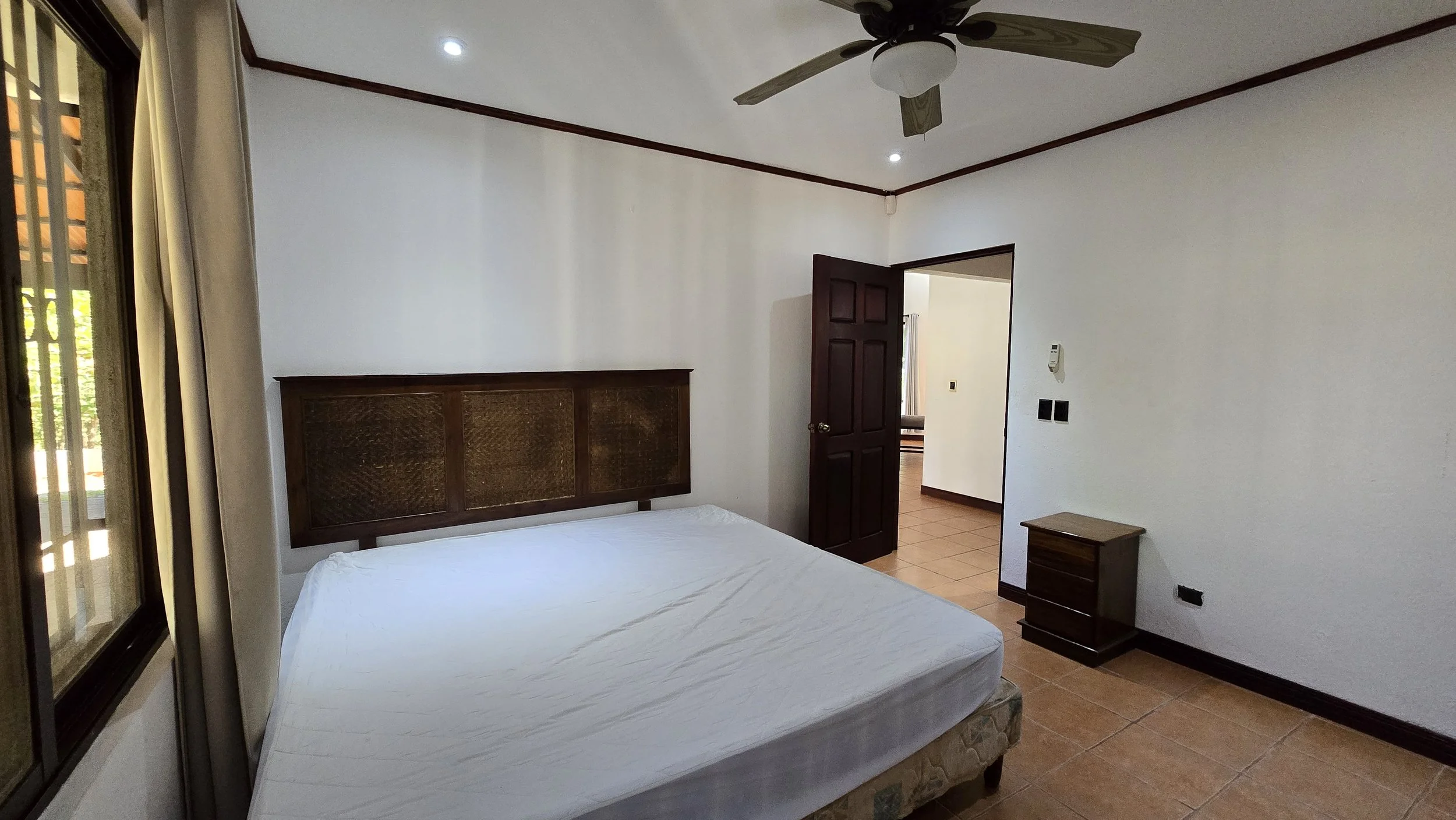 Empty bedroom with a white bed, wooden headboard, small wooden nightstand, ceiling fan, window with curtains, and an open door leading to another room.