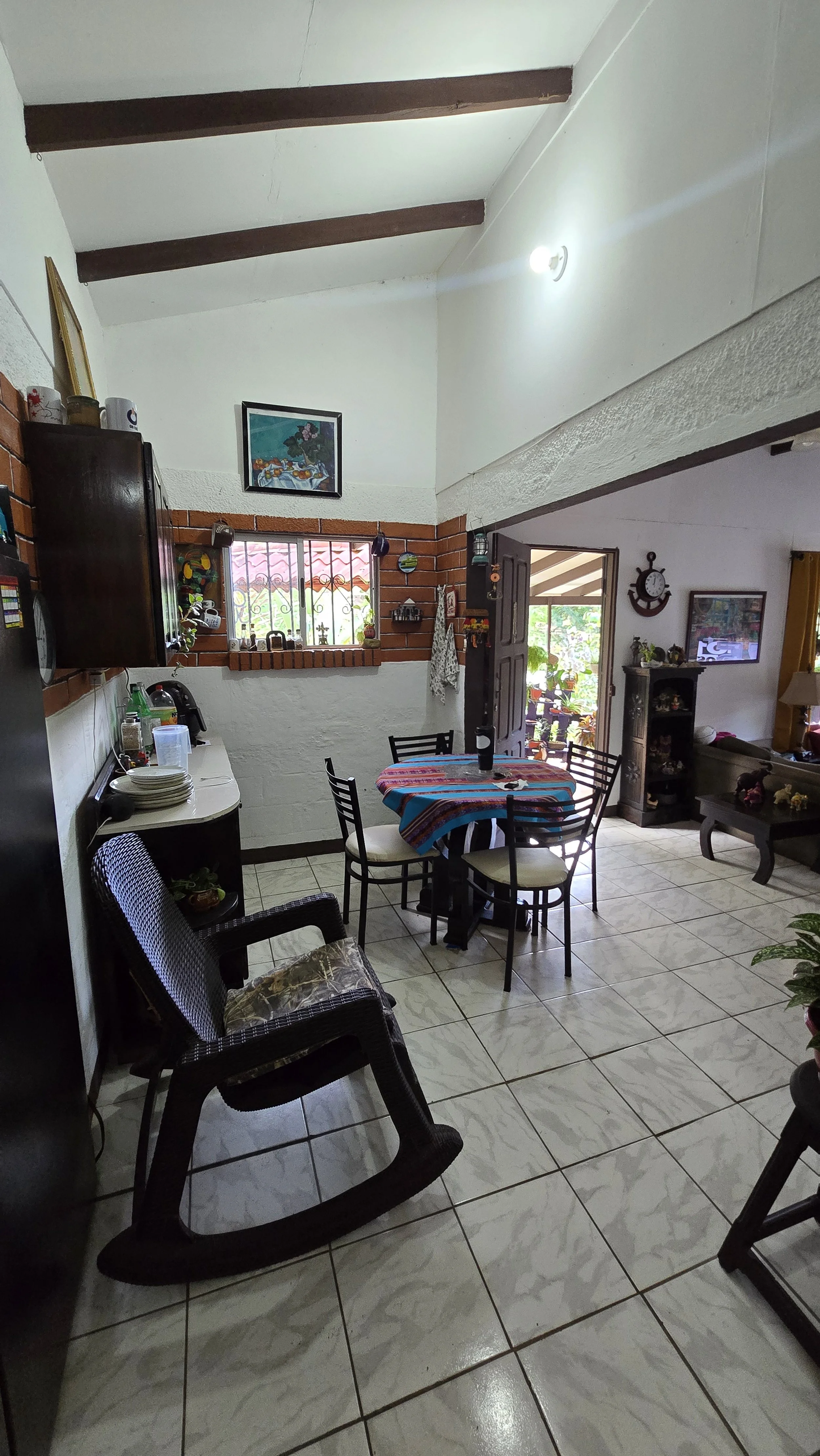 Interior view of a dining area with a round table covered with a colorful striped tablecloth, surrounded by five black chairs with white cushions. There is a black rocking chair with a cushion near the table, and a small corner cabinet with dishes an