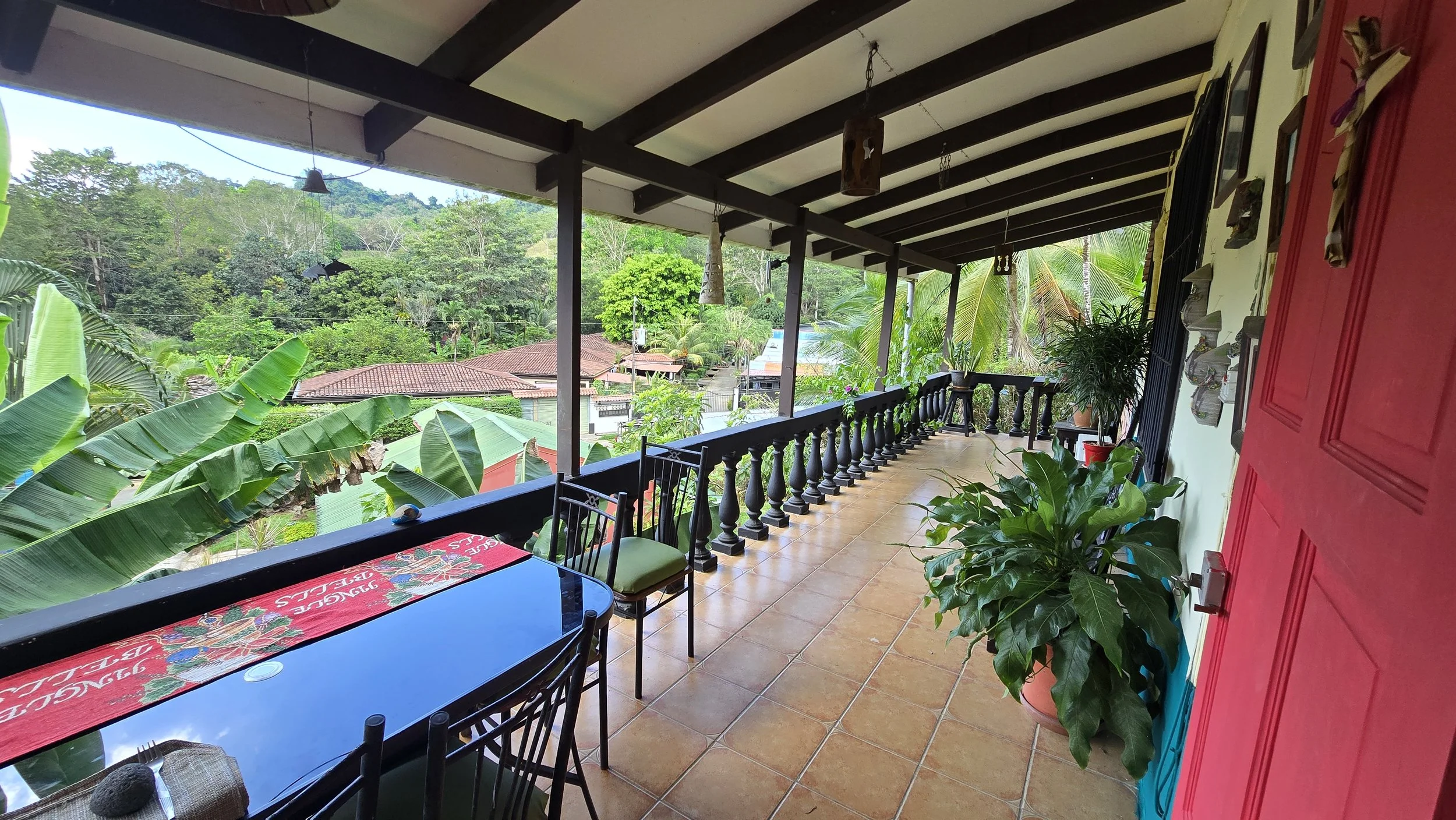 A balcony with a tiled floor, black railing, and ceiling with exposed beams. There are potted plants along the railing and a red door on the right. A table with a red cloth and chairs are in the foreground. Green trees and rooftops of houses are visi