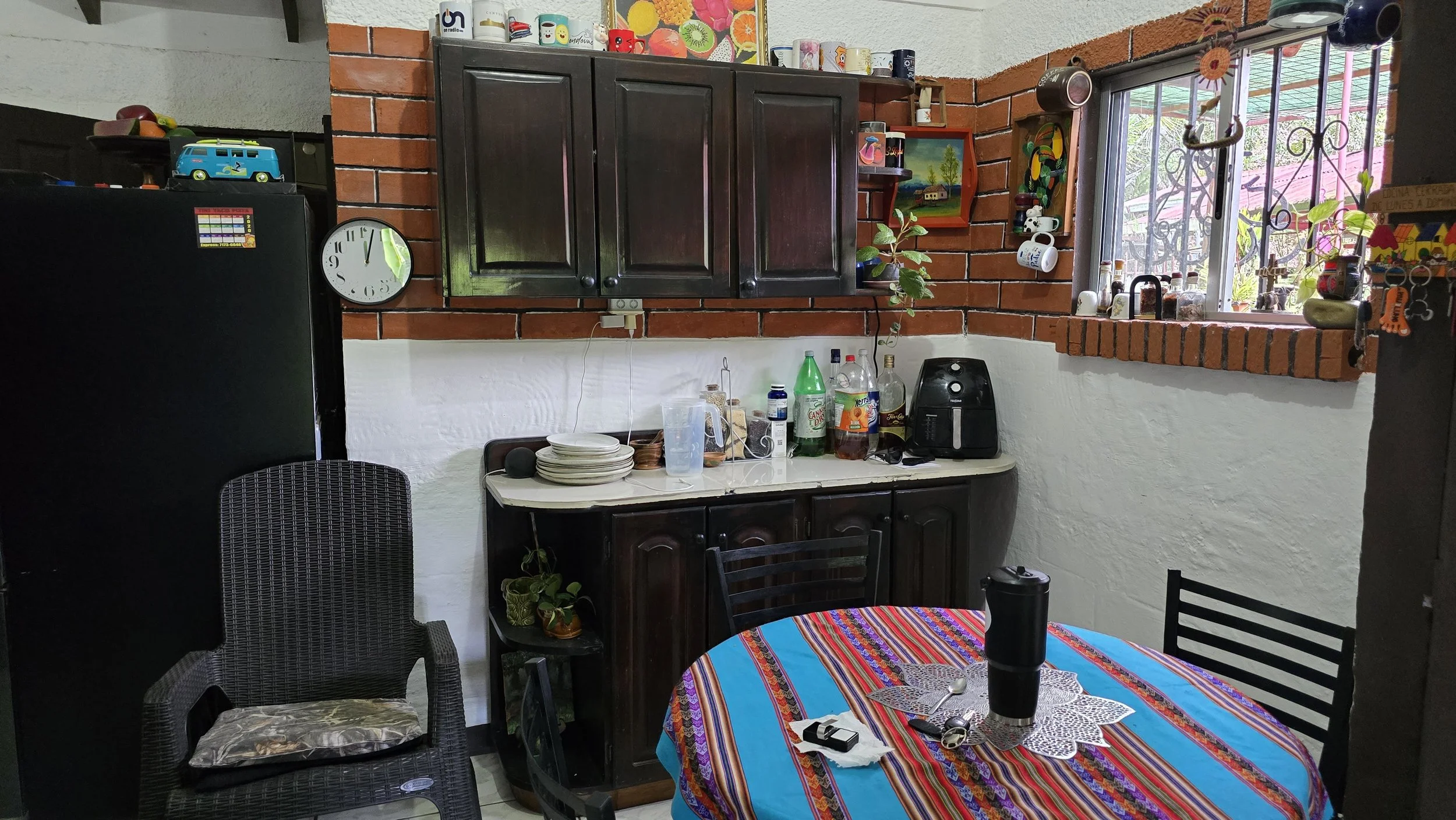 A kitchen with a small round table covered with a colorful striped tablecloth, surrounded by black chairs, and various hidden items on the table, including keys and a thermos. The backdrop features a white wall with a black refrigerator on the left, 