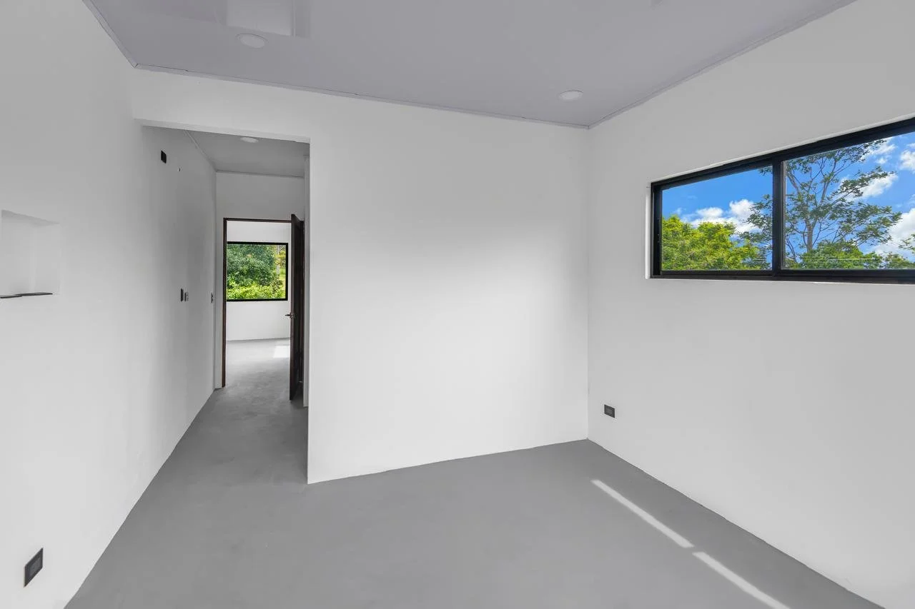 Empty white room with large window showing green trees and blue sky, doorway leading to another room, gray carpeted floor, and minimal ceiling lights.