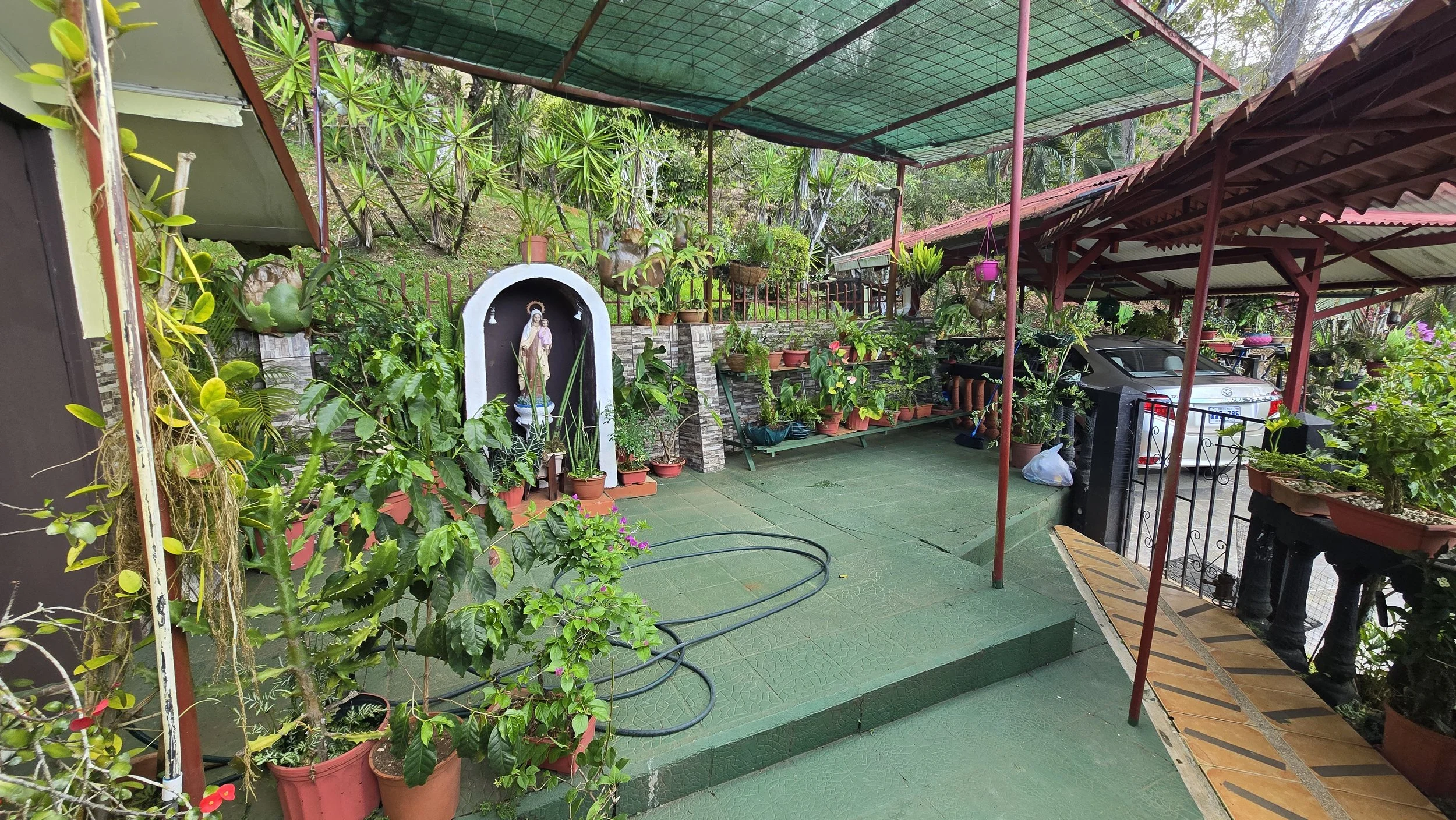 A covered patio area filled with numerous potted plants and flowers, with a religious statue in a small shrine, and a car parked under a roof on the right side.