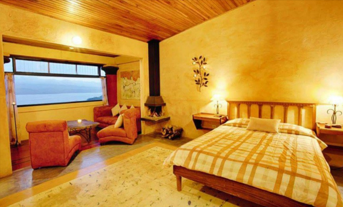 Cozy bedroom with a large bed, nightstands with lamps, a sitting area with pink armchairs, a window with a view, and a fireplace with logs.