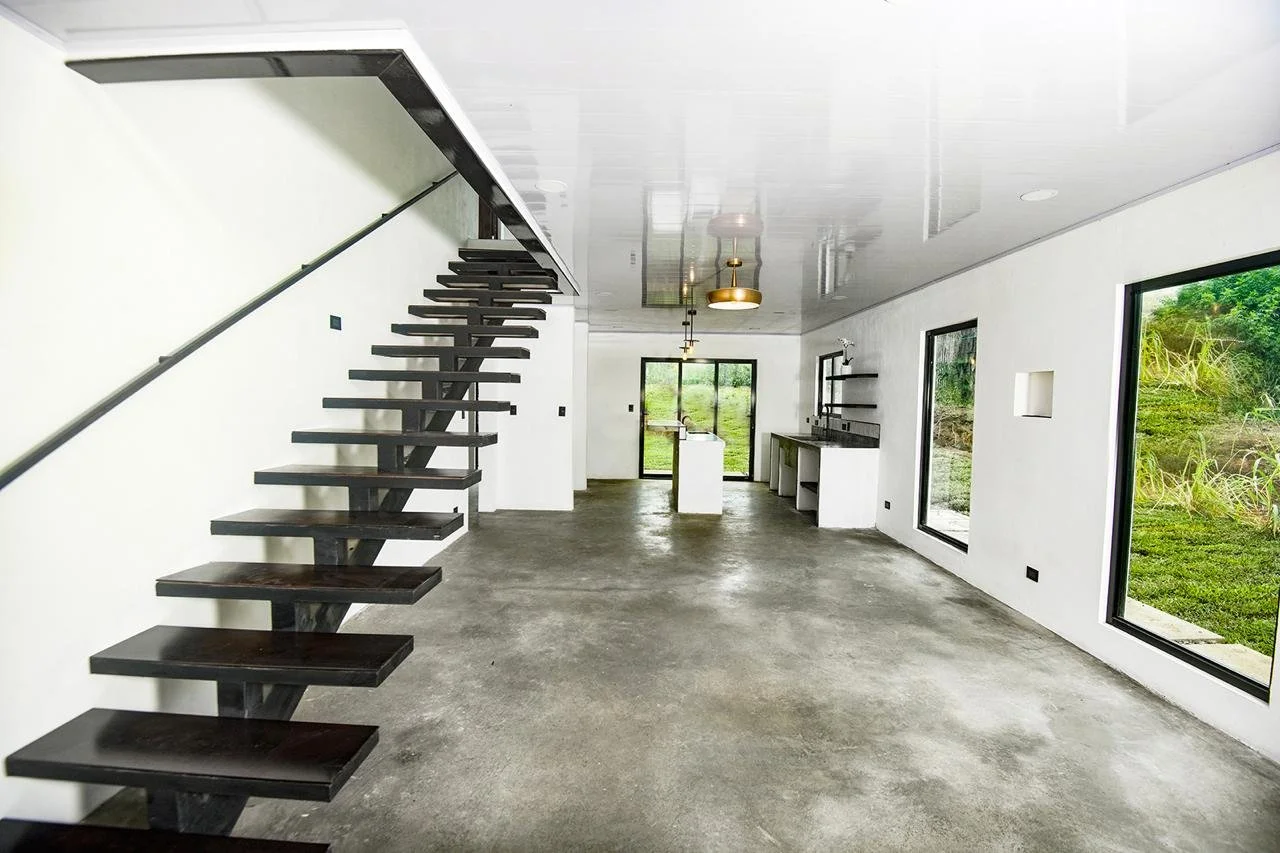 Empty modern living space with concrete floor, white walls, large windows, and a staircase with dark wooden steps and black railing.