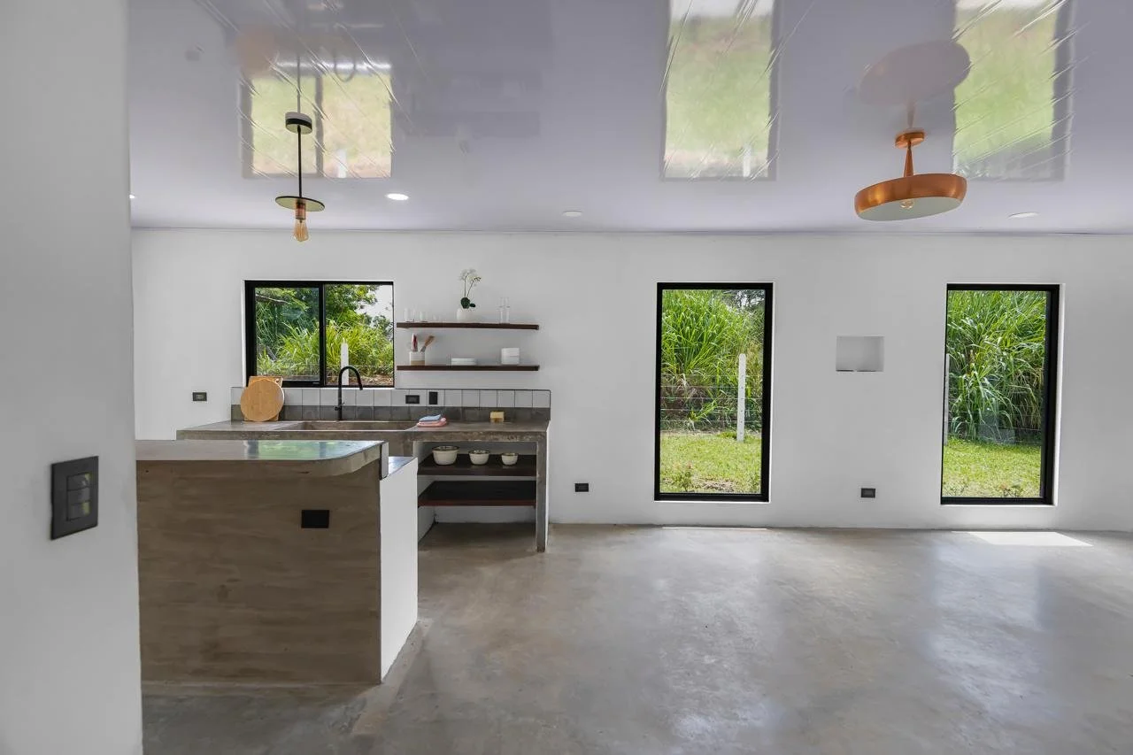 Minimalist kitchen with white walls, black framed windows showing green outdoor foliage, a small countertop with a sink, open wall shelves, and two ceiling fans, one black and one copper.