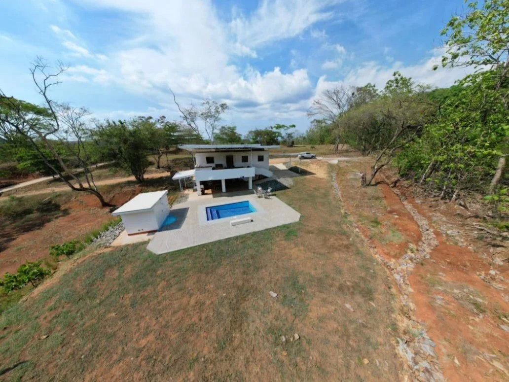 A modern house with a swimming pool and a small detached structure in the backyard, surrounded by trees and a dirt pathway on a partly cloudy day.