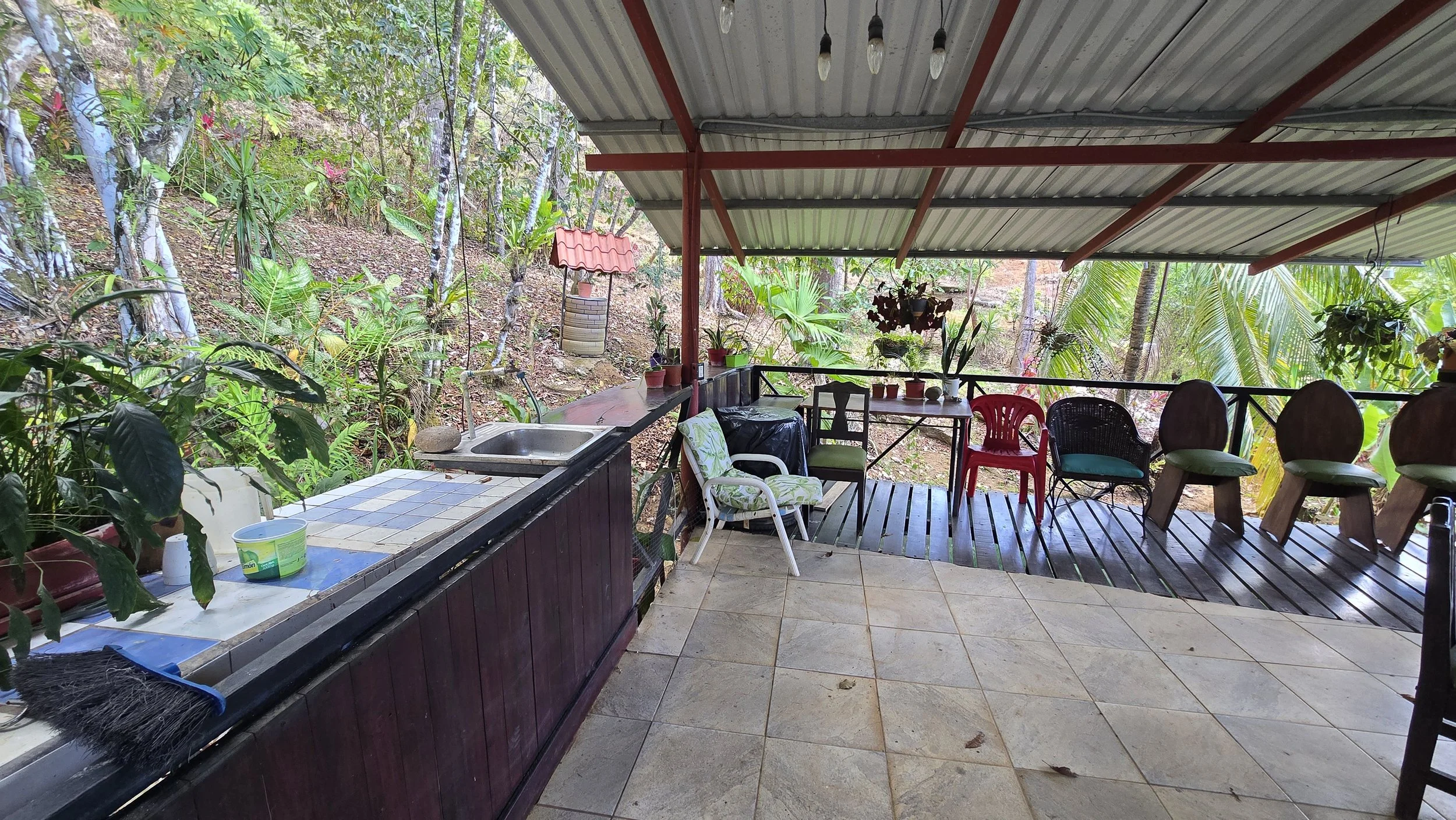 Open outdoor porch with tiled and wooden flooring, surrounded by lush tropical vegetation, with hanging plants and a small well with a red roof in the background.