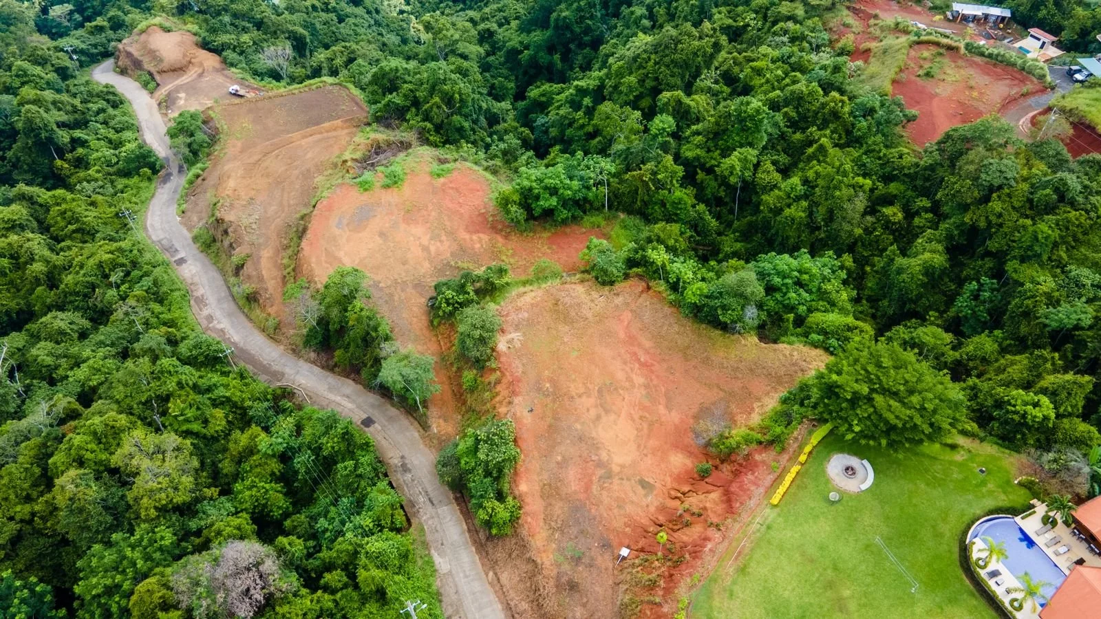 Aerial view of a partially cleared hillside surrounded by dense green trees, with a winding road along the left side and a residential property with a lawn, pool, and a small structure in the bottom right corner.