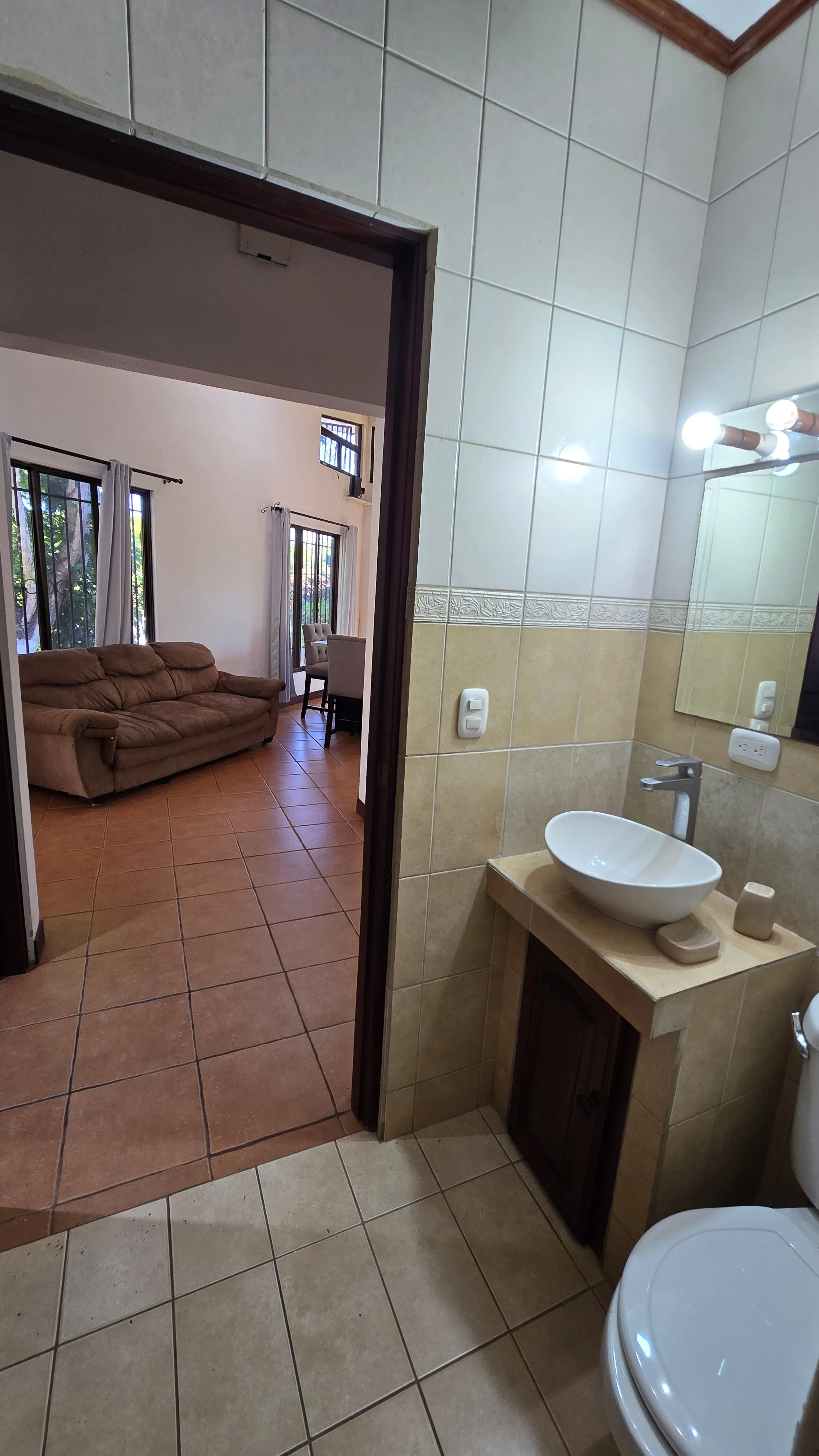 Bathroom with beige tiled walls and floor, small wooden cabinet with a vessel sink, mirror, and light fixtures. Visible part of a toilet. Adjoining living room with tiled floor, beige sofa, and windows with curtains.