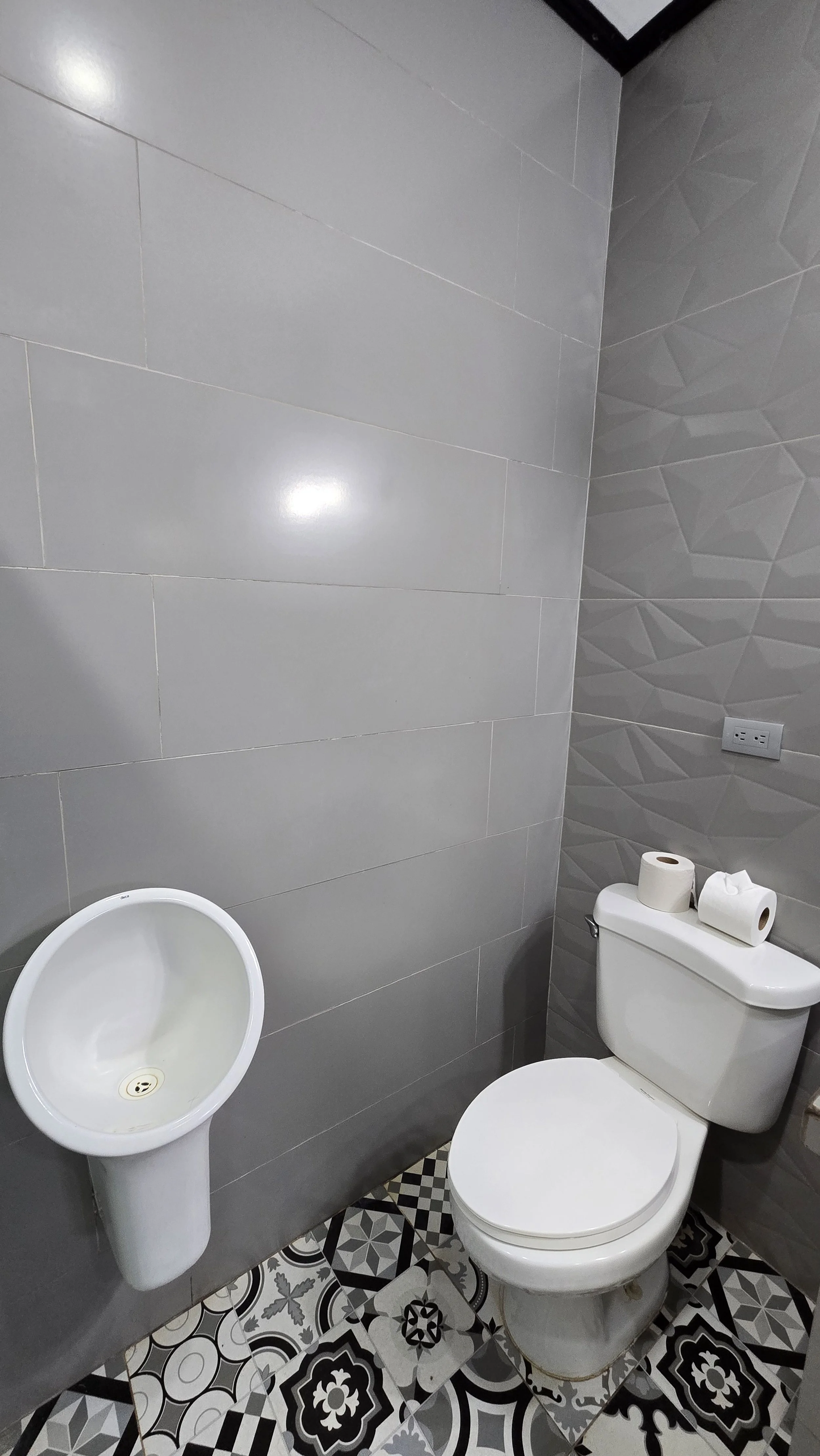 A small bathroom with a white toilet, two rolls of toilet paper on the tank, a urinal on the wall, gray tiled walls, and patterned black and white floor tiles.