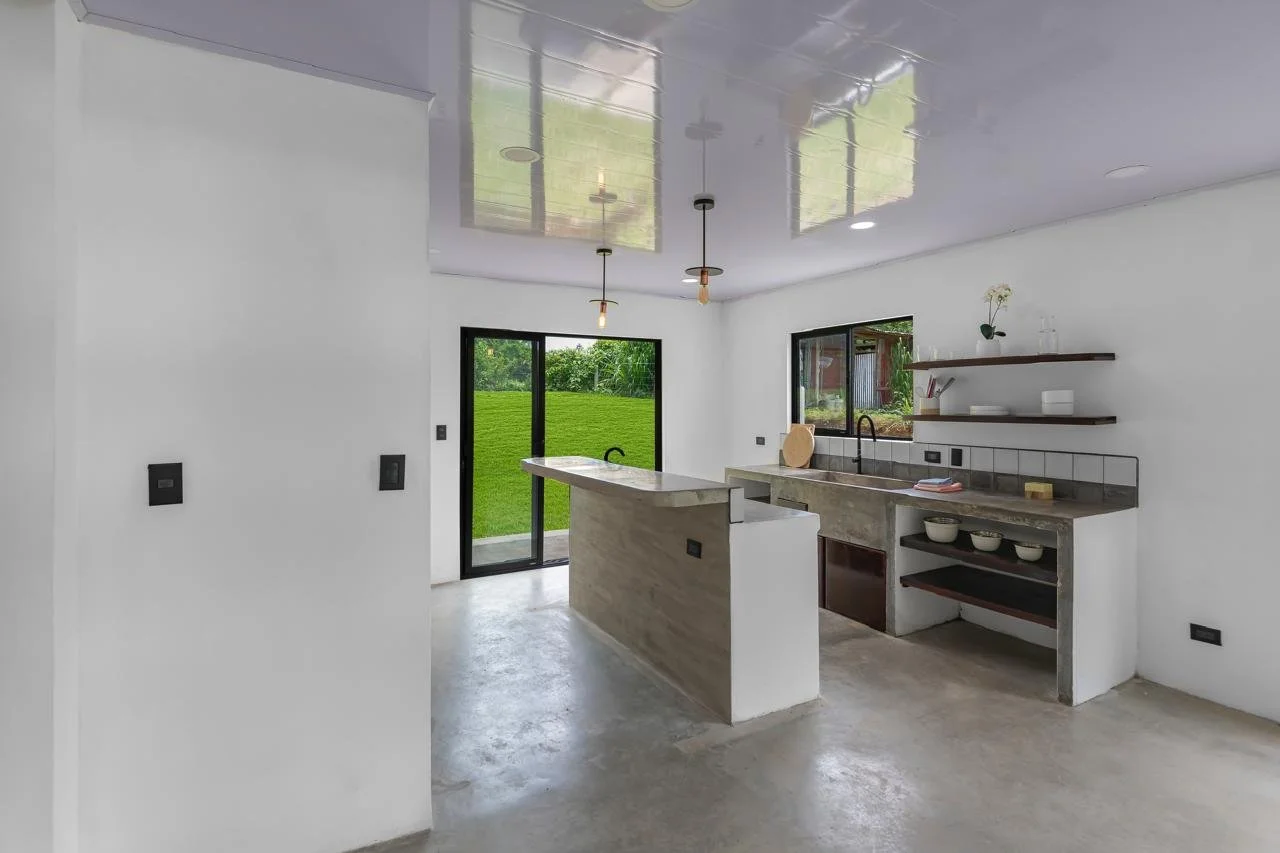 Modern kitchen with white walls, concrete floor, and black window and door frames. Features a central island with a marble countertop, a sink with a faucet, and open shelving with bowls and decorative items. Two hanging light fixtures and a sliding g