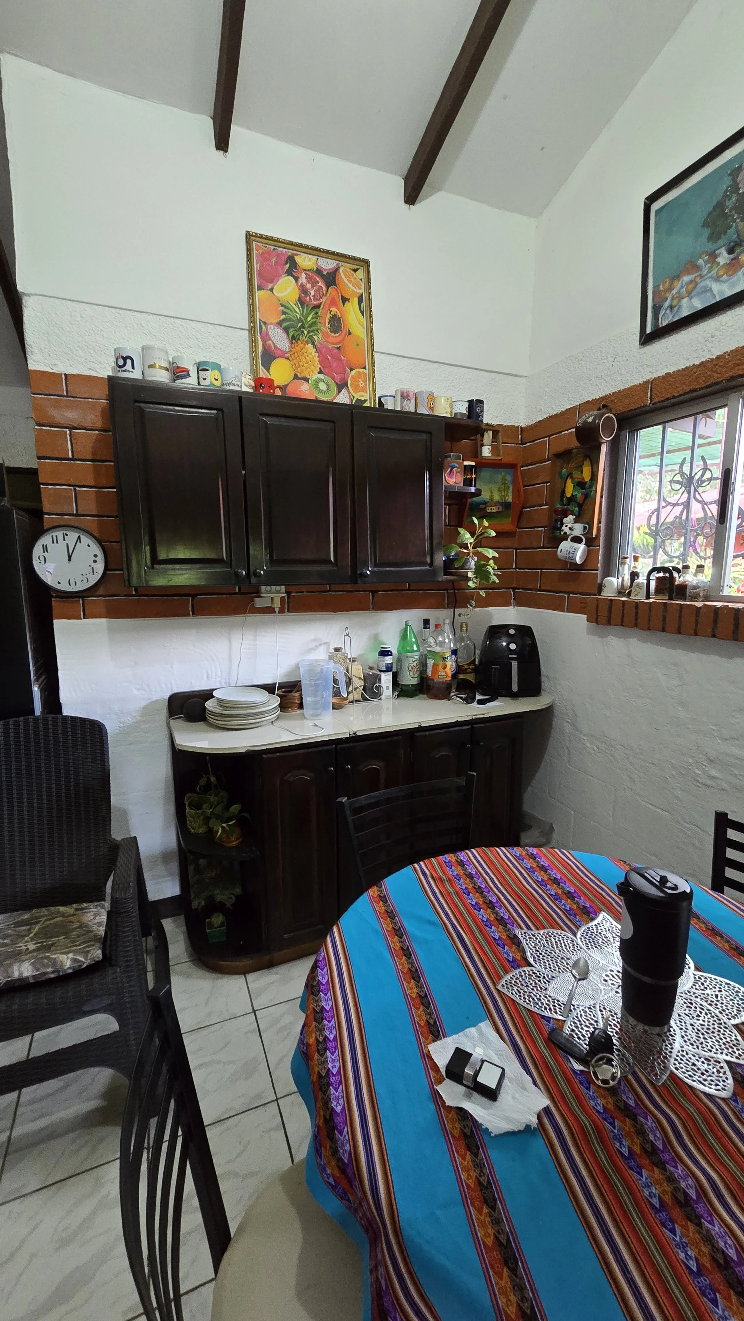 A dining area with a round table covered with a colorful striped tablecloth, black chairs, a black cabinet with a white countertop holding bottles, cups, and dishes, a window with decorative ironwork, and wall art including a framed fruit picture.