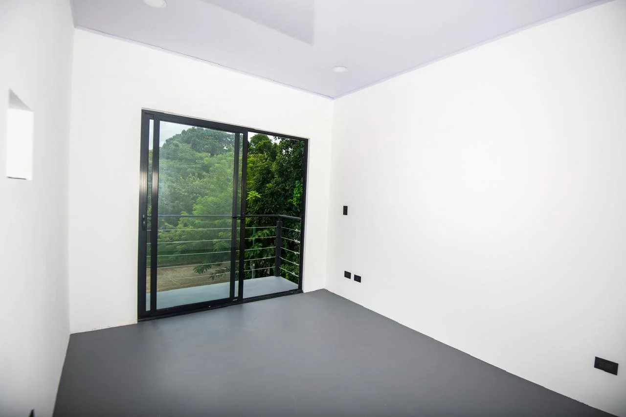 Empty room with gray flooring, white walls, and a sliding glass door leading to a balcony with a view of green trees outside.