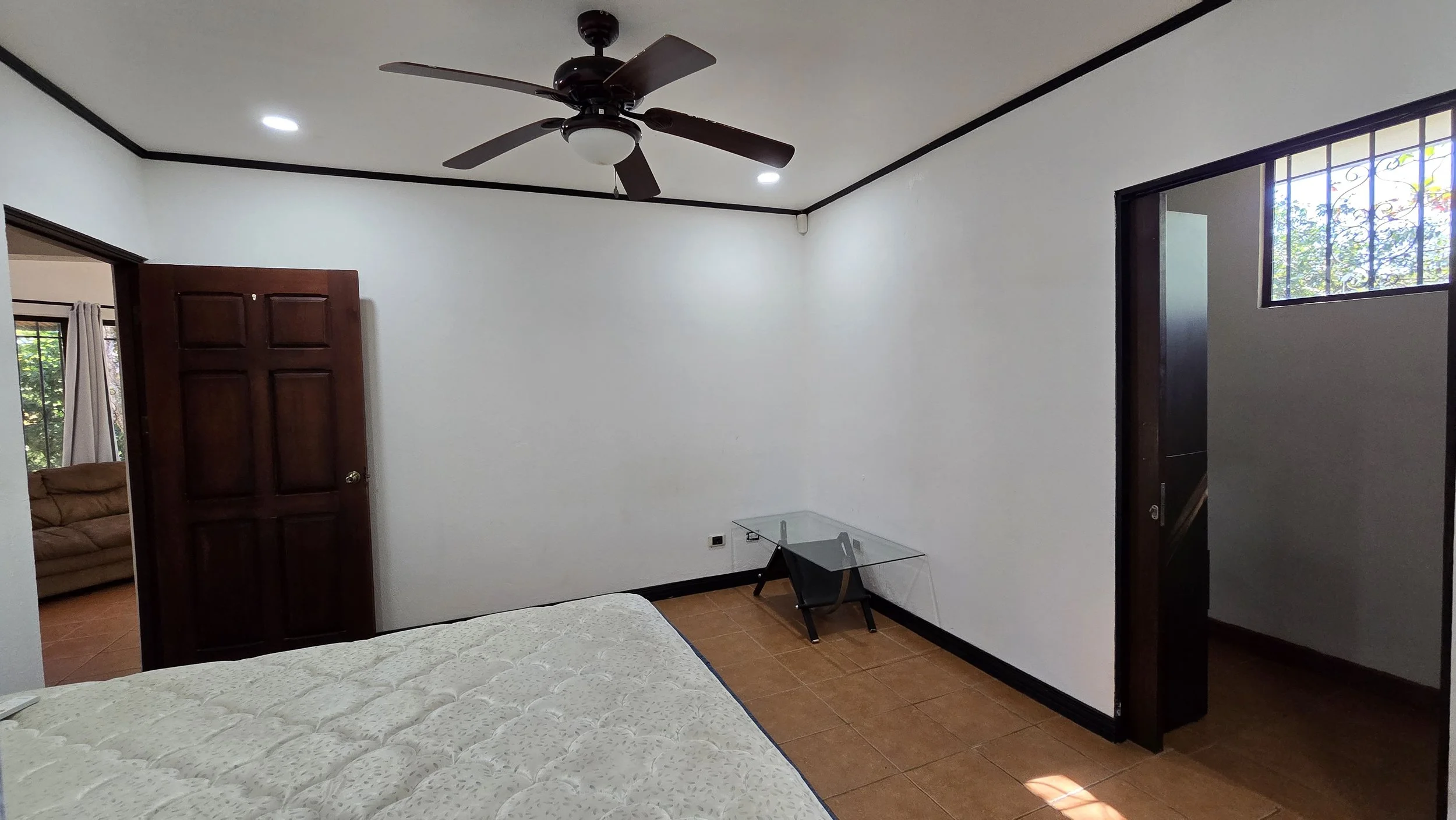 Empty bedroom with white walls, black trim, wooden door, small glass desk, and ceiling fan.