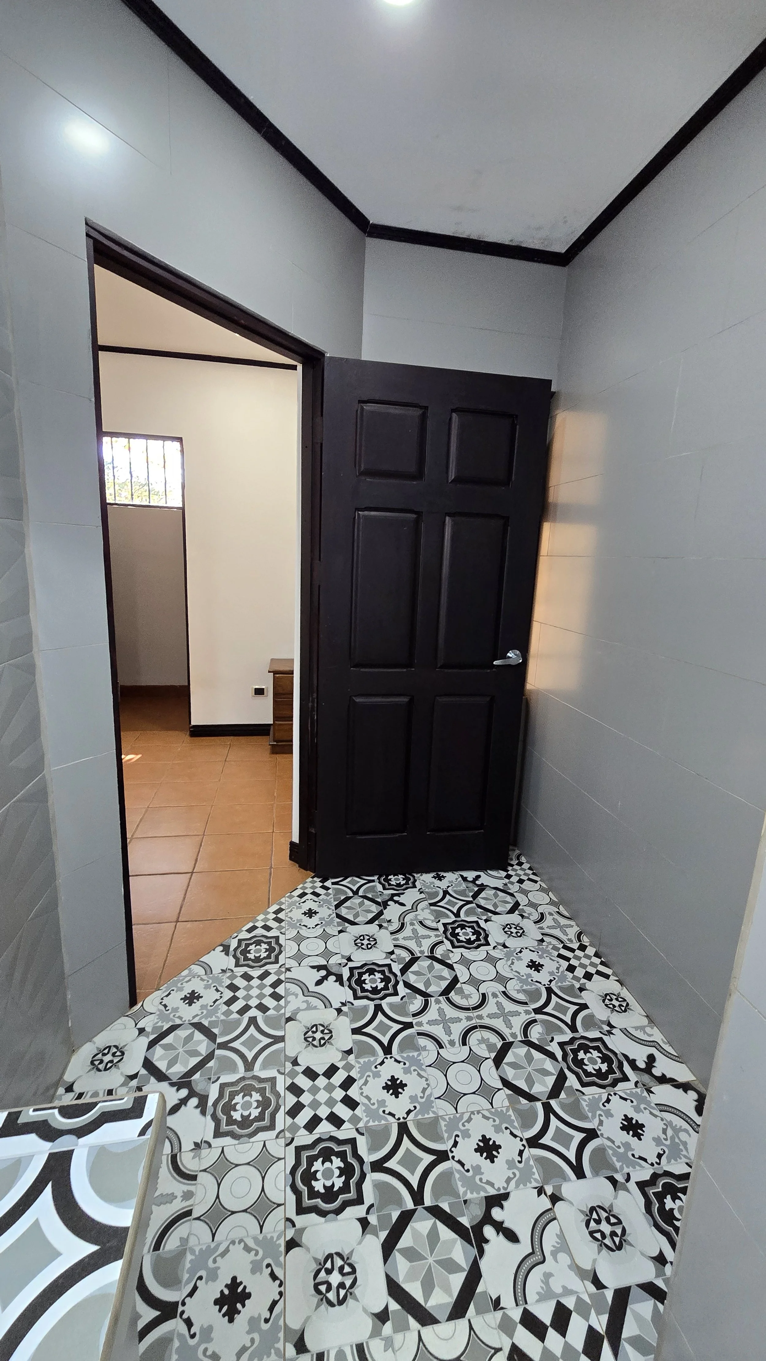 A doorway with a black door slightly open, revealing a room with tiled flooring and a small window with metal bars, and a glimpse of a wooden piece of furniture.