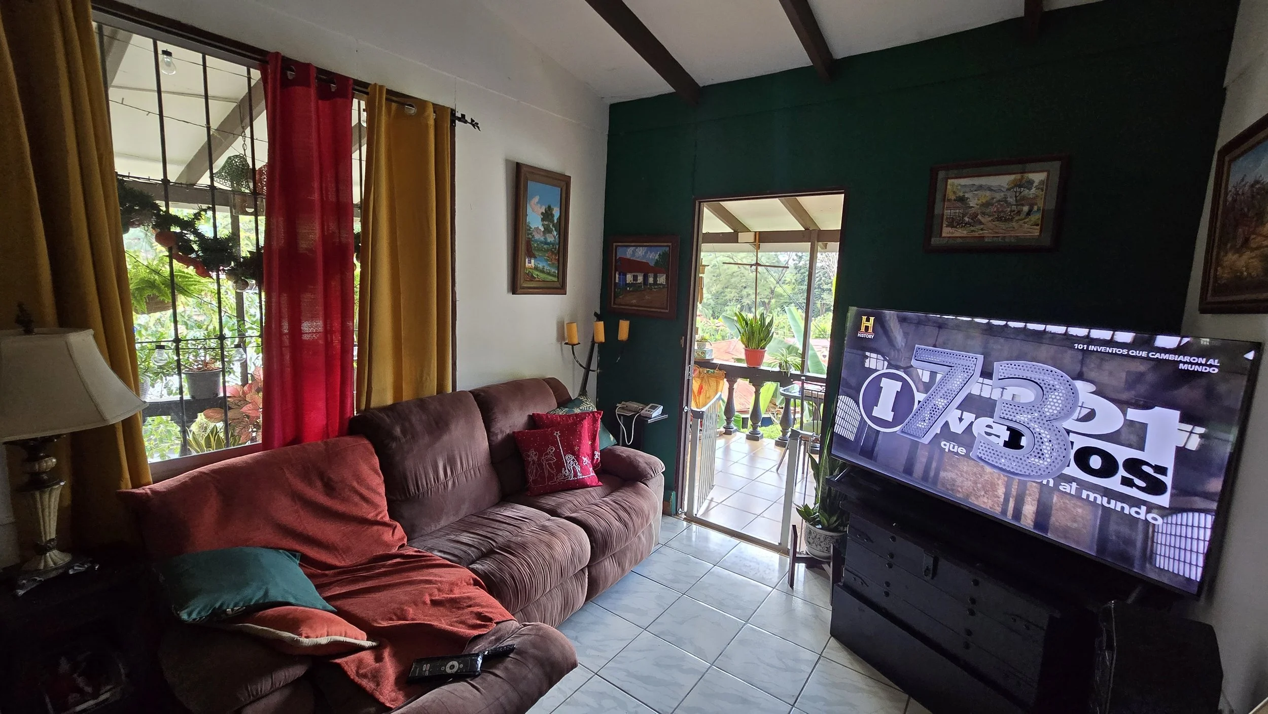 Living room with a brown sofa, colorful pillows, and a television showing a program. There are large windows with red and yellow curtains, a balcony with plants outside, and framed paintings on the wall.