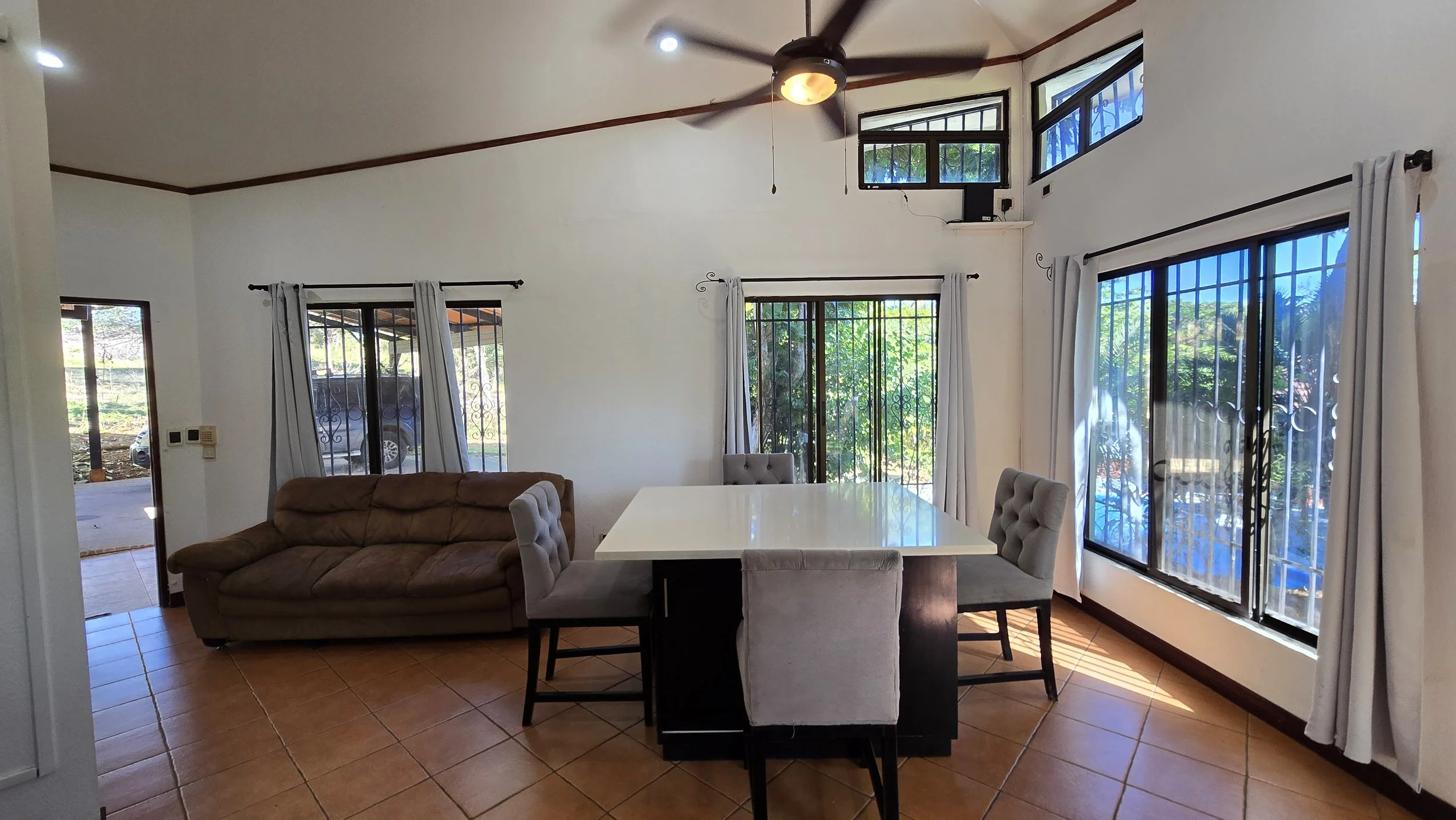 Living room with a brown sofa, a white dining table, and four upholstered chairs. Large windows with white curtains, a ceiling fan, and natural light.