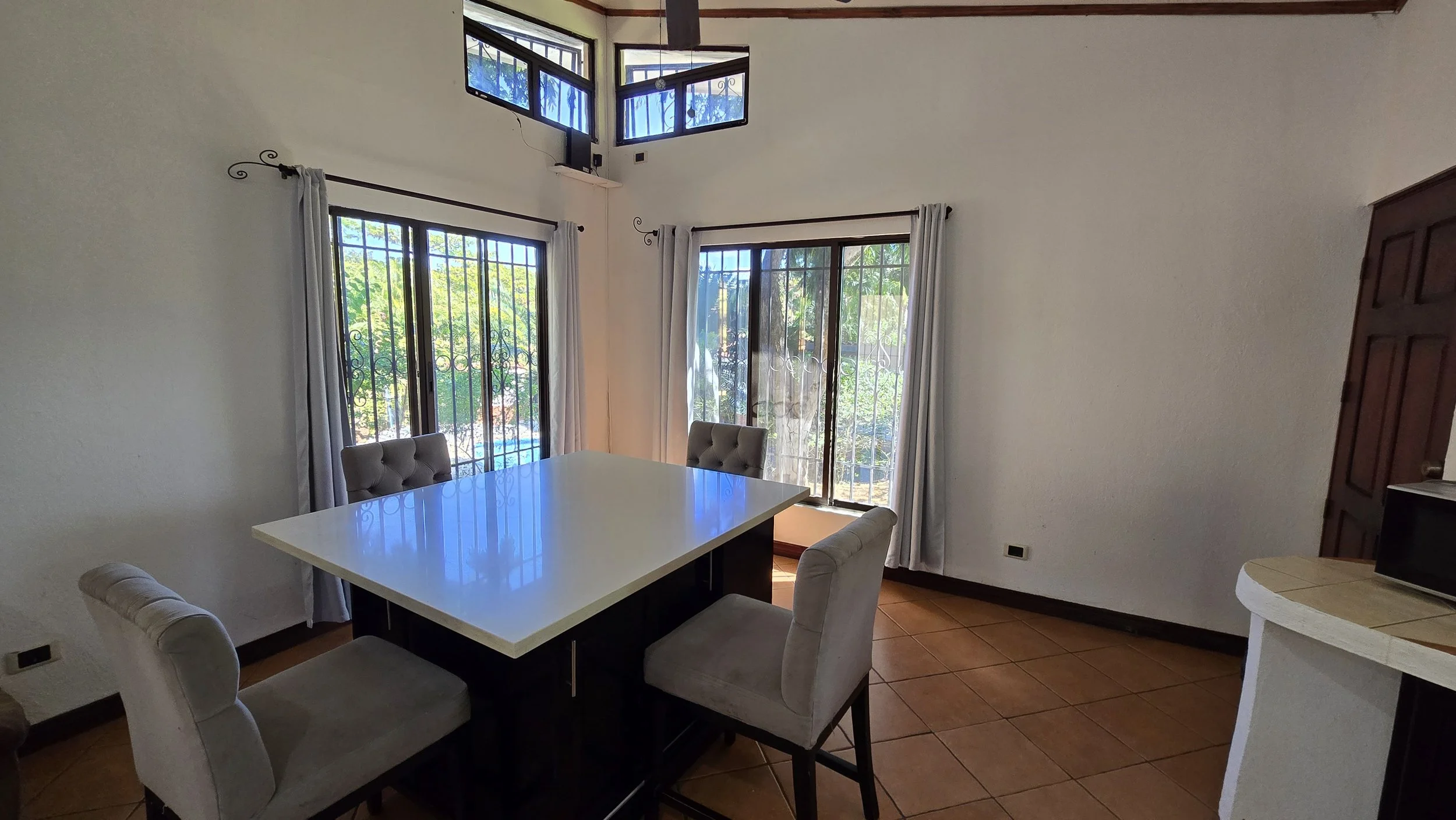 Dining area with a white table, four upholstered chairs, large windows with security bars, white curtains, tiled floor, and a high ceiling with corner windows.