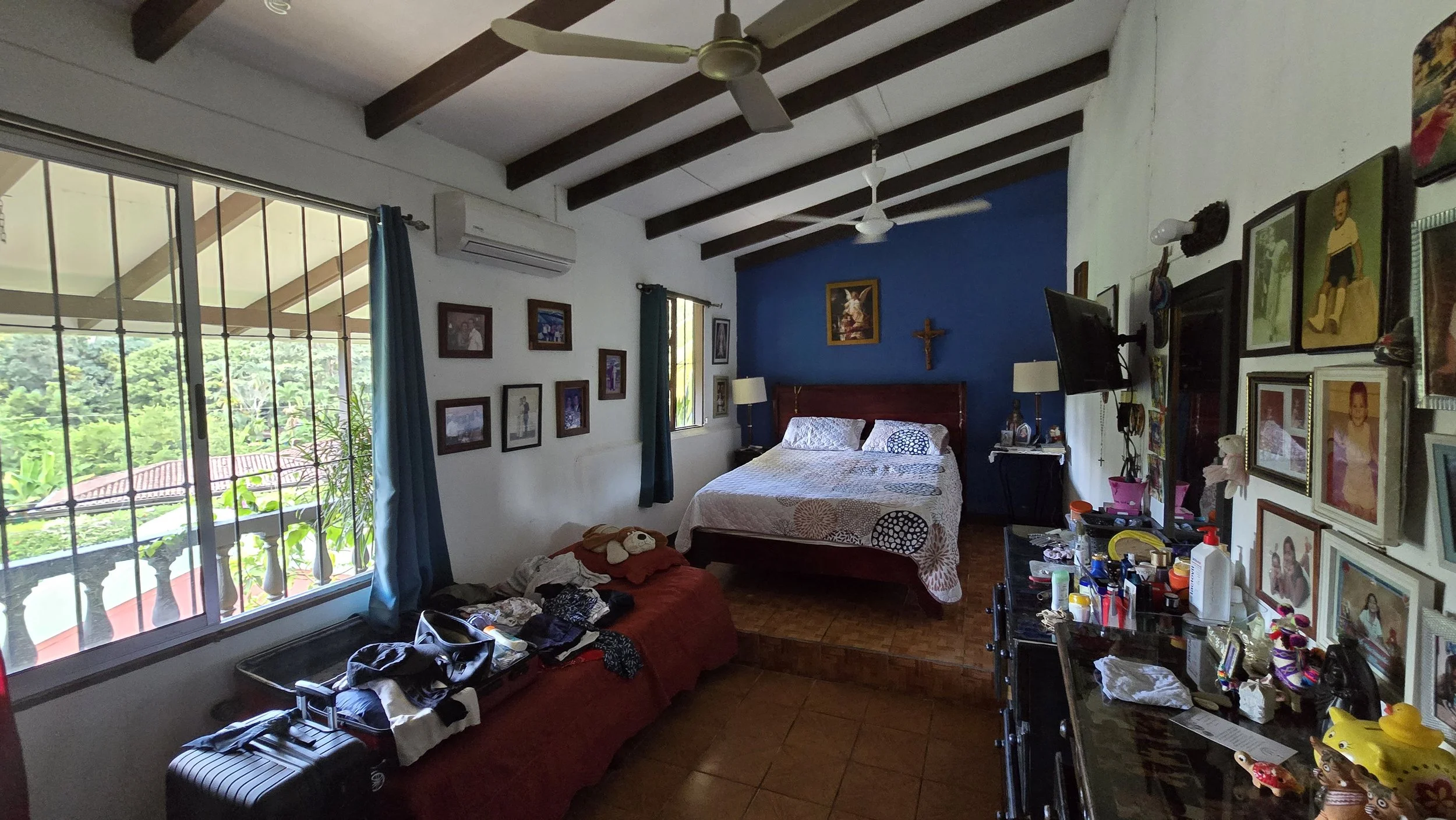 A cozy bedroom with a large window showing greenery outside. The room features a bed against a blue accent wall with religious decor, including a crucifix and religious pictures. There is a ceiling fan, a collection of framed photos on the wall, and 