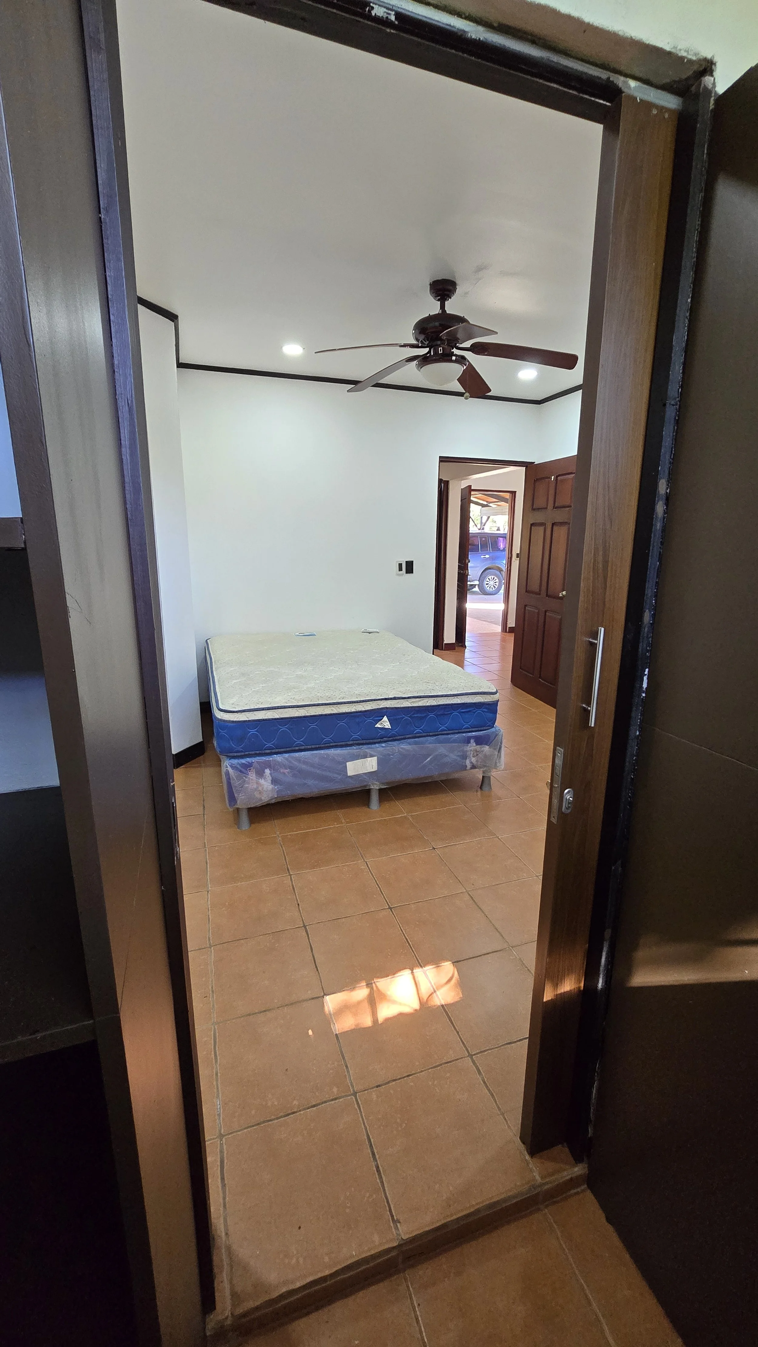 Empty bedroom with a mattress on a bed frame, brown tiled floor, ceiling fan, and an open door leading outside.