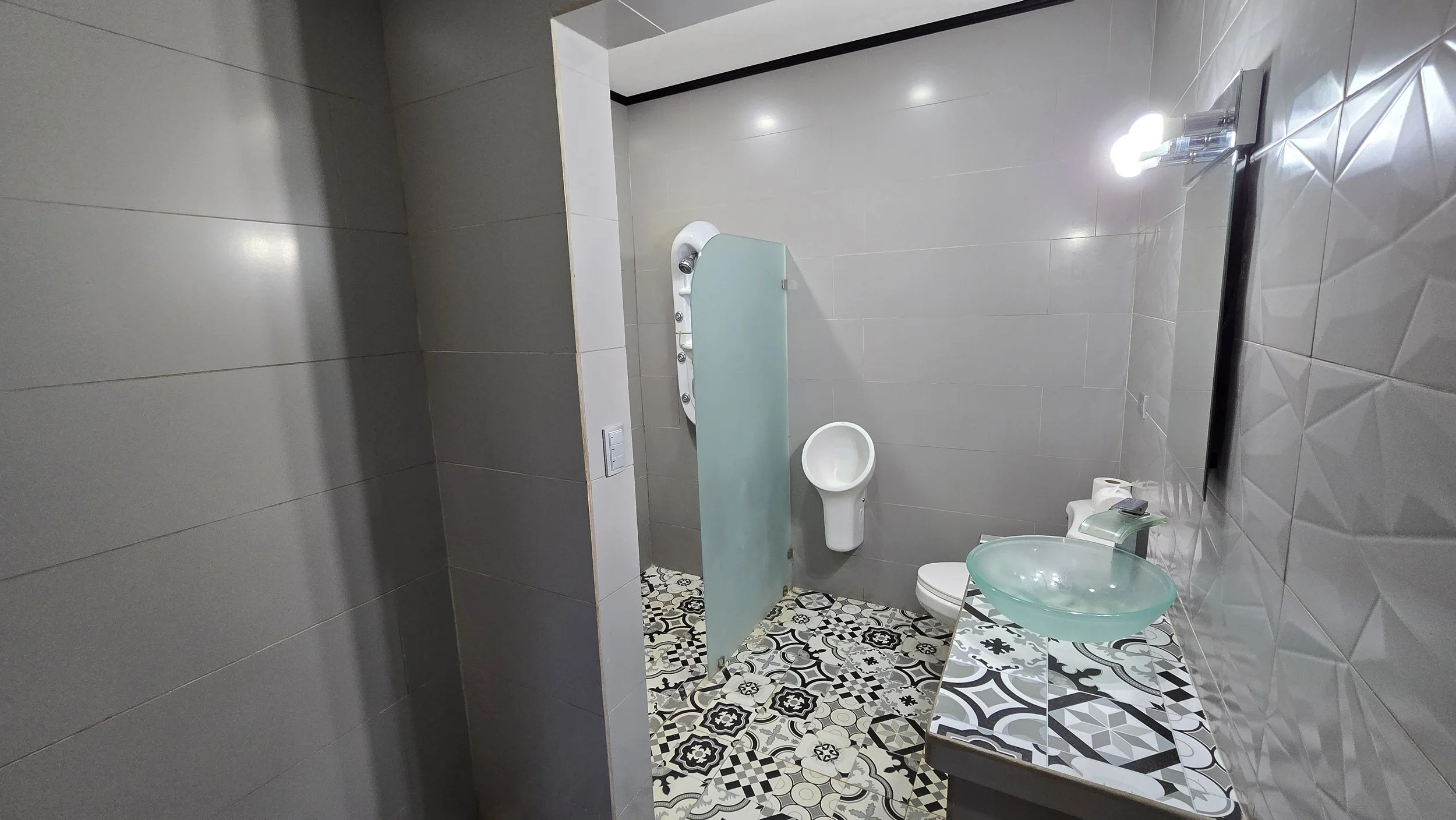 Interior view of a modern bathroom with patterned floor tiles, a frosted glass vessel sink on a tiled countertop, a wall-mounted mirror, toilet paper, a urinal, a privacy partition, and a wall-mounted TV.
