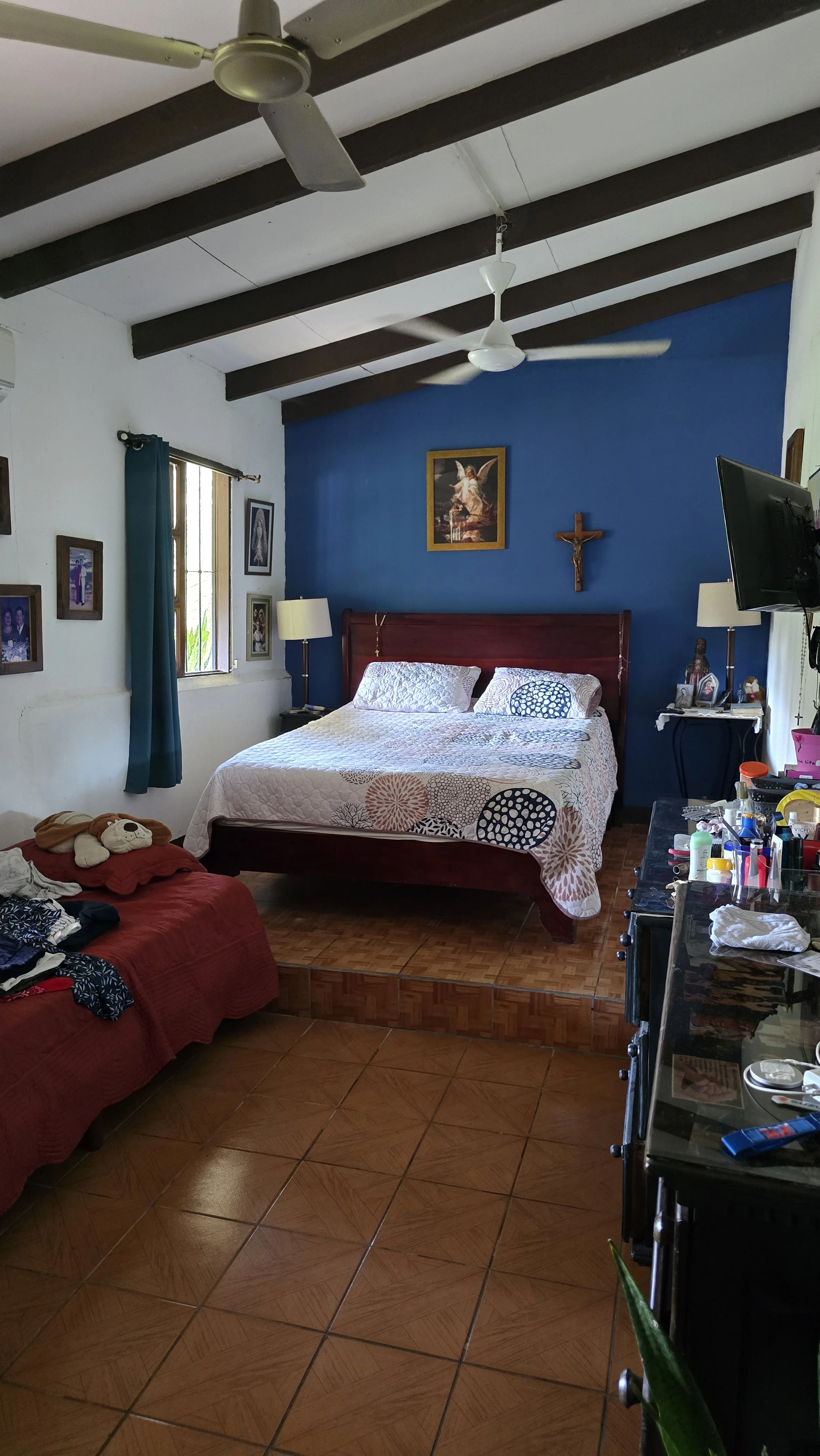 A bedroom with a white ceiling, dark wooden beams, and ceiling fans. There is a bed with a wooden headboard against a blue accent wall, decorated with religious pictures and a crucifix. To the left, there are windows with blue curtains, and a red cou