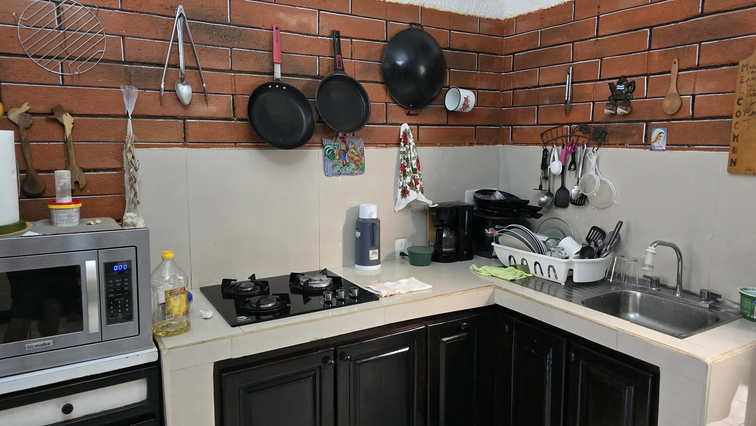 Kitchen countertop with stove, microwave, coffee maker, dishes, hanging pots, pans, and cooking utensils on brick and tiled wall.