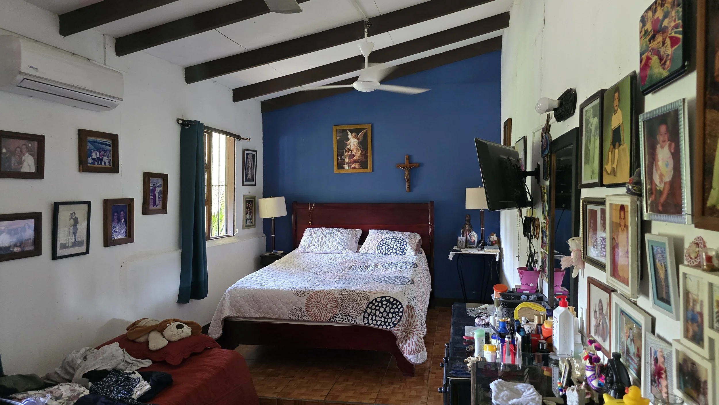 A bedroom with a bed against a blue accent wall, a crucifix and a religious painting above it. The room has a ceiling fan, a window with green curtains, multiple family photos on the walls, a bedside table with a lamp, a flat-screen TV, and various p