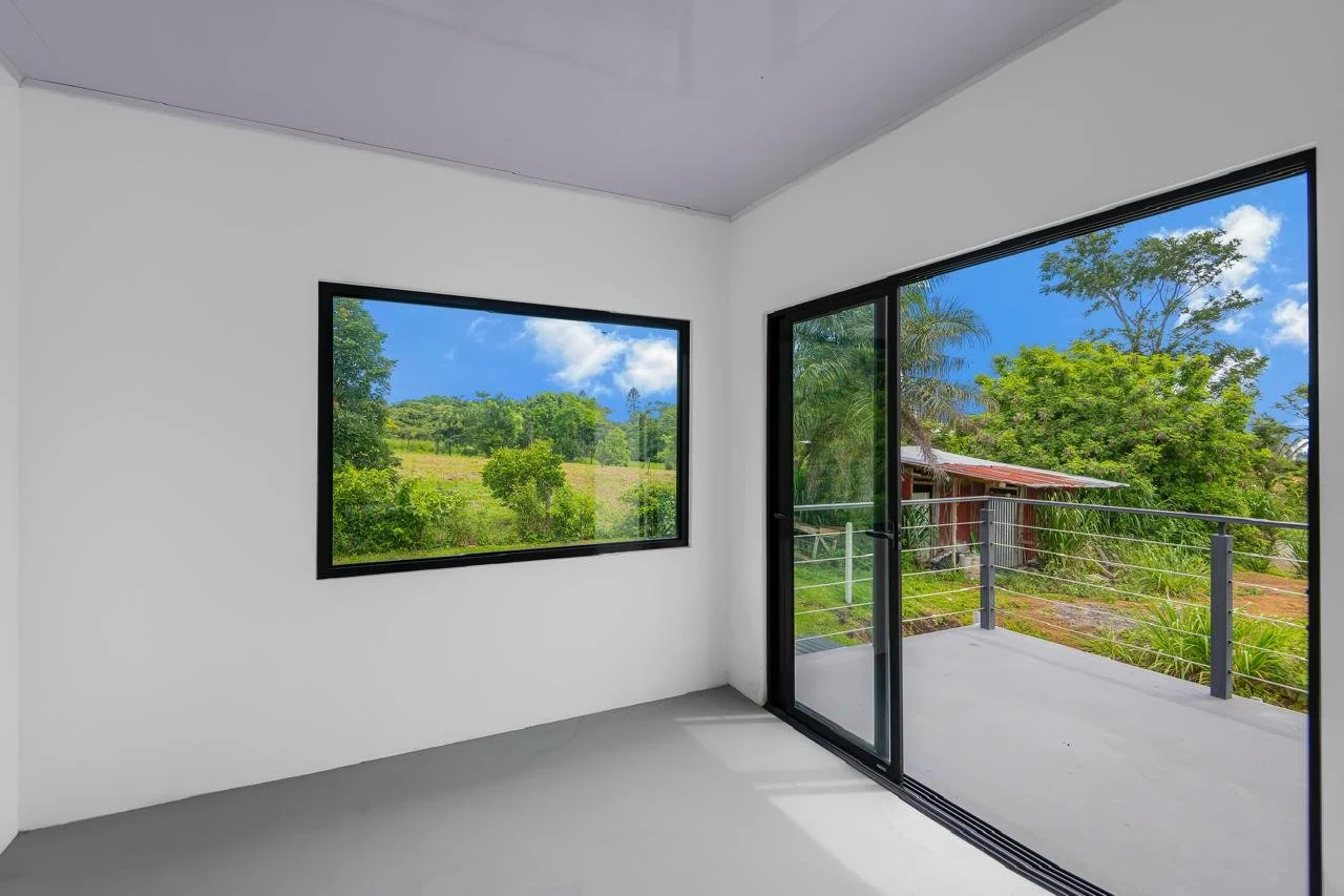Empty room with two large windows showing a scenic outdoor view of green trees, plants, and a blue sky with clouds, and a sliding glass door leading to a balcony with a metal railing.
