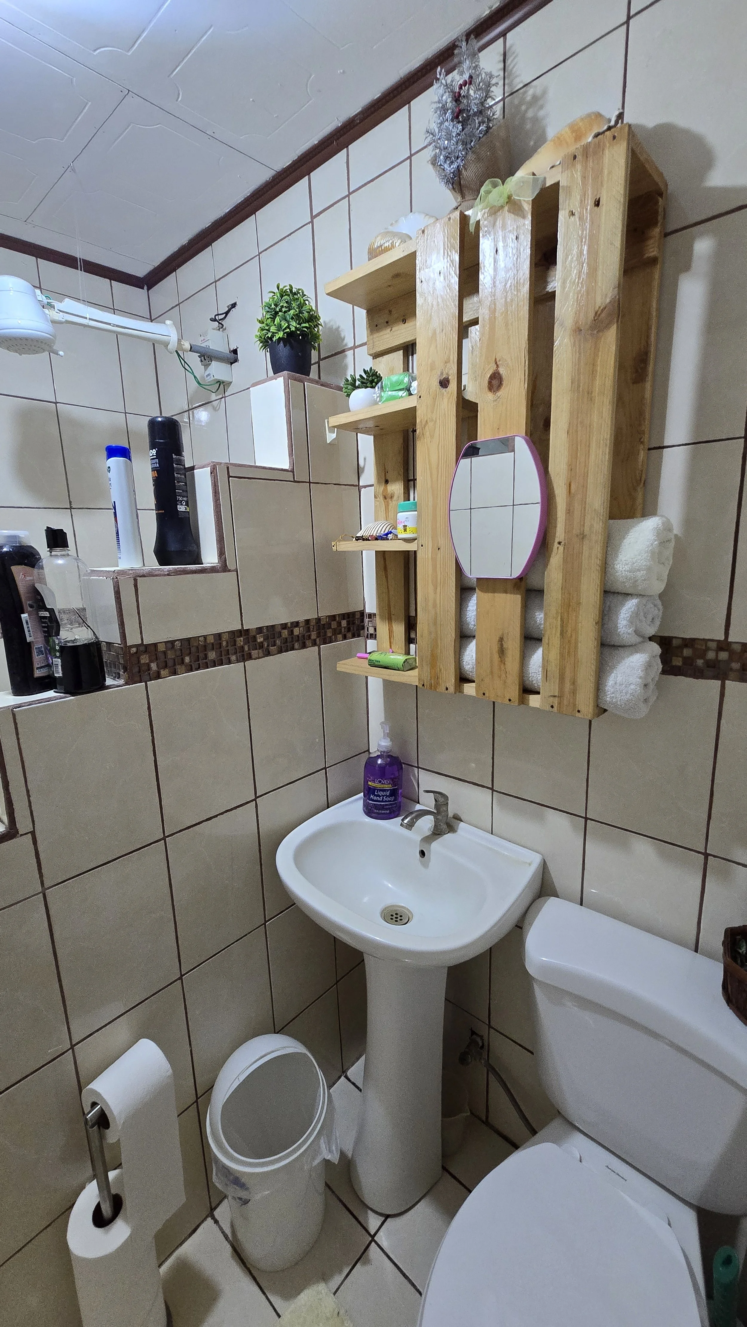 Small bathroom with beige tiled walls, white pedestal sink, toilet, and small trash can. Wooden pallet used as a shelf above the sink holds rolled towels, toiletries, and decorative items. There is a mirror on the pallet and a hand soap pump on the s