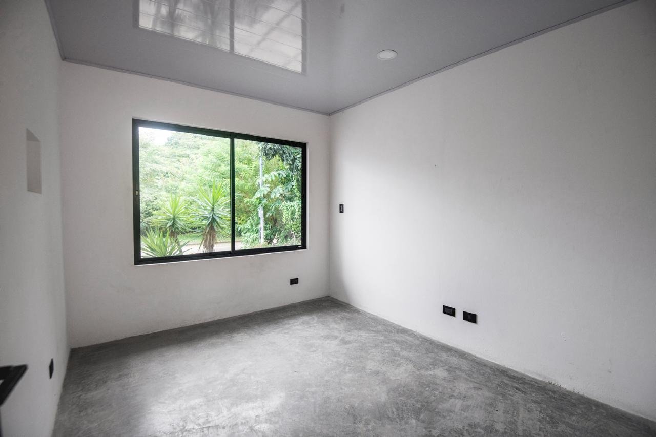 An empty room with a large window showing green trees outside, plain white walls, and a concrete floor.