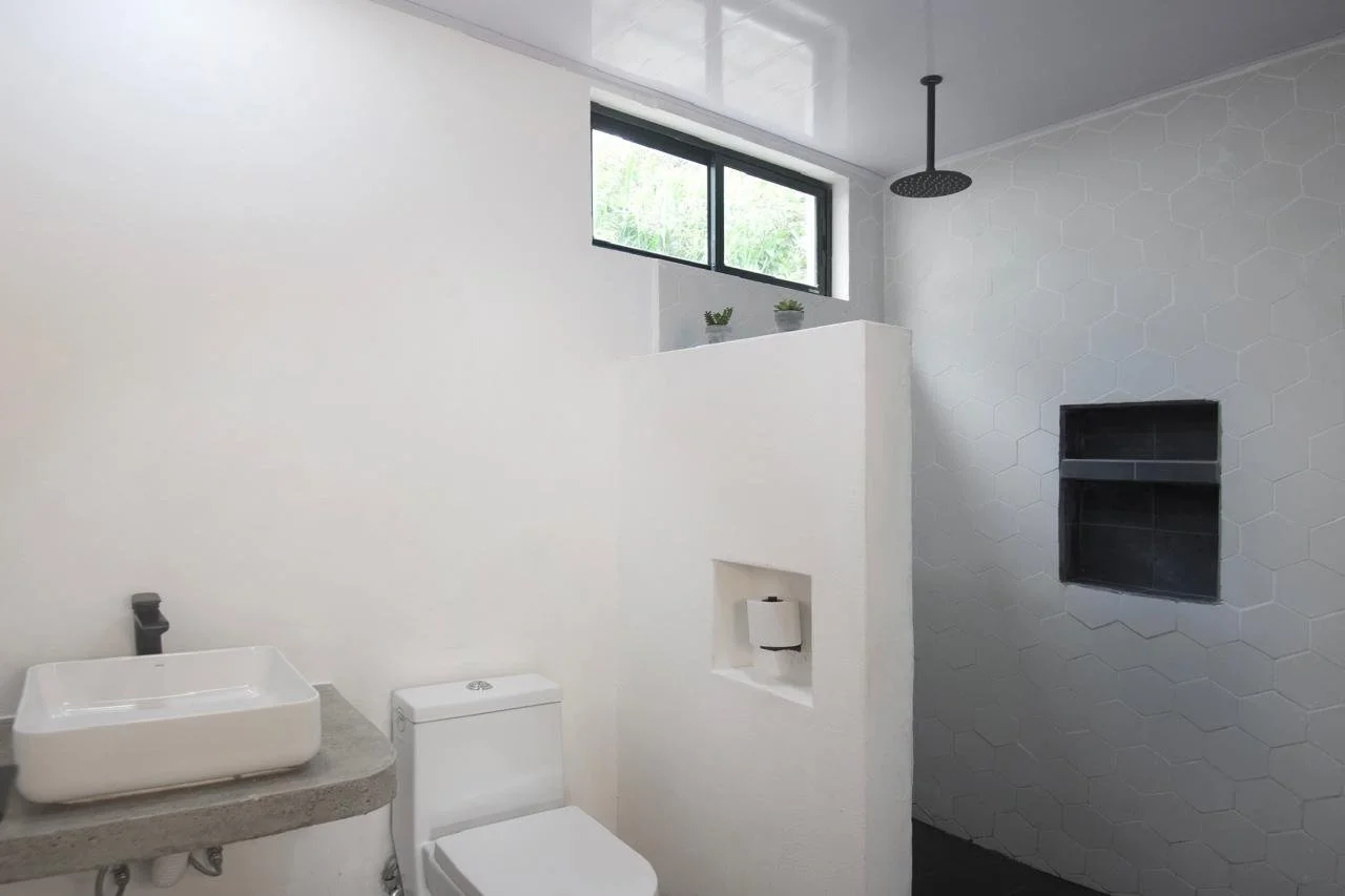 Modern bathroom with white walls, a concrete sink, a white toilet, a large window, two small plants, and a black showerhead.