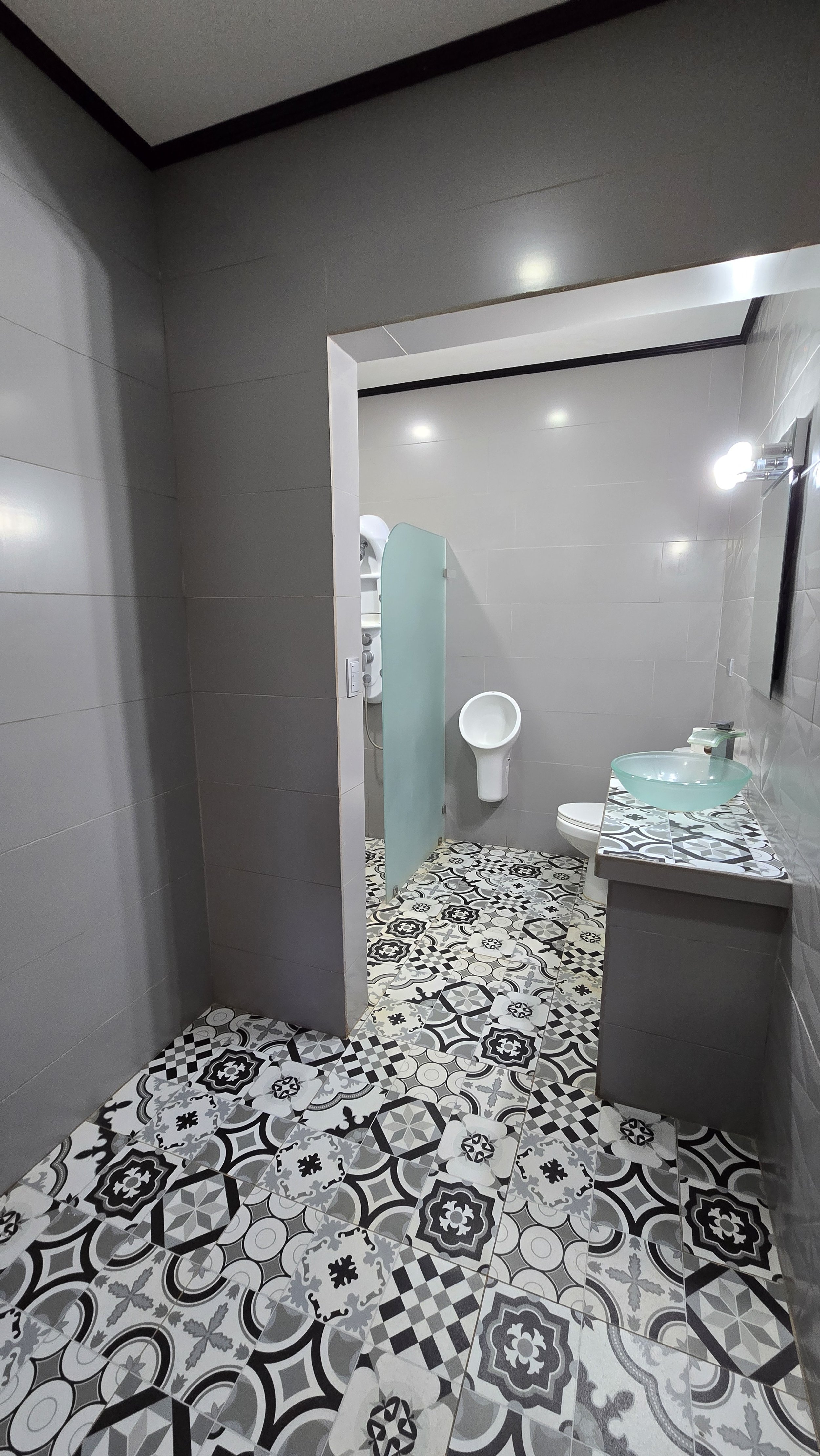 A modern public restroom with patterned tile floor, a hand-washing sink with a glass basin, a mirrored mirror, a wall-mounted lamp, a toilet, and a urinal separated by a frosted glass partition.
